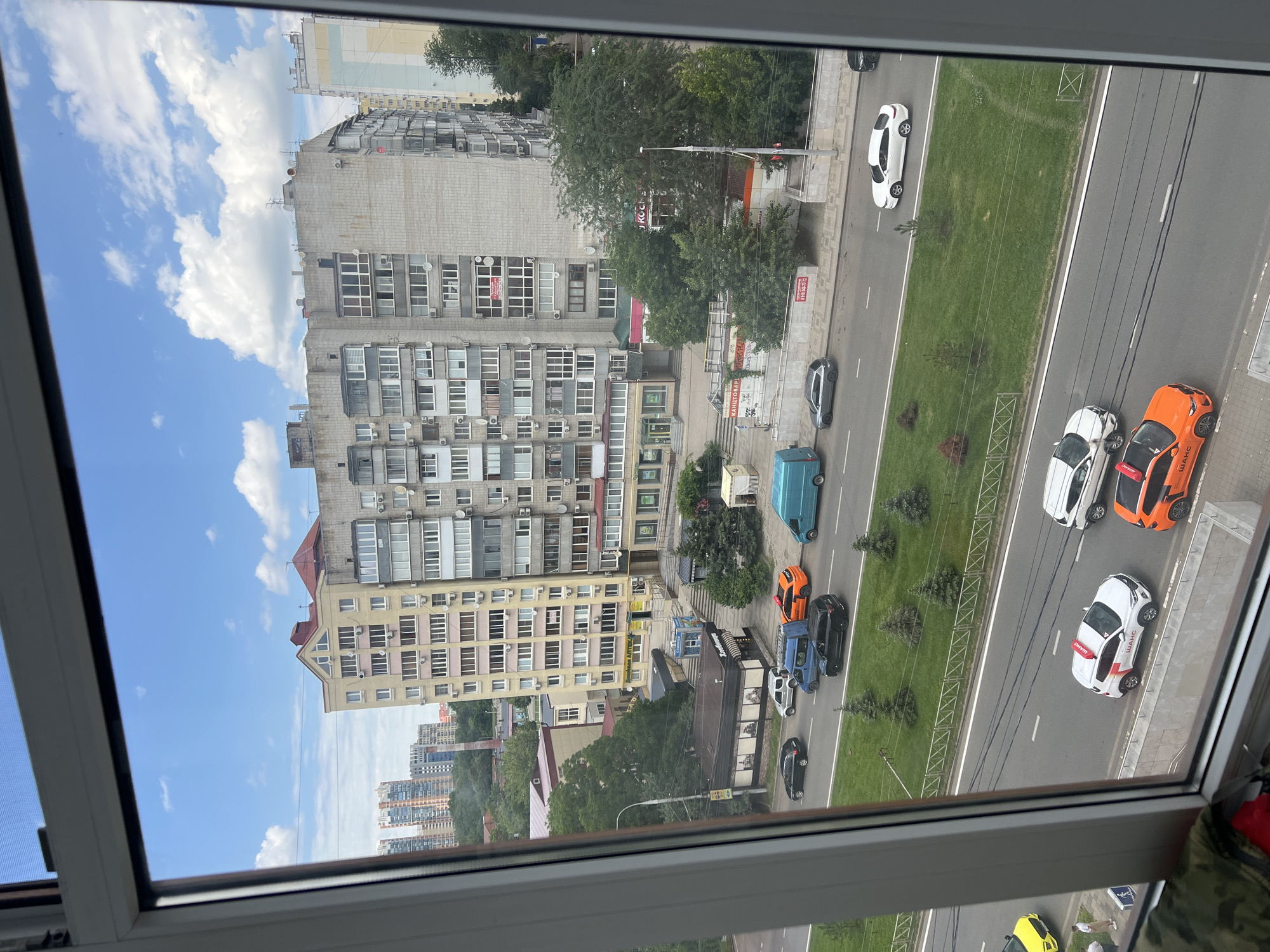 A medium shot captured from a high vantage point through a window shows a city street scene under a partly cloudy sky. The view is framed by the white window frame on the left and right edges of the image.
The street is lined with buildings of varying heights and architectural styles. On the left, a taller, light-yellow building features multiple floors with balconies and windows, some with pink accents around the windows. To its right, a larger, grey multi-story building dominates the mid-ground, characterized by its concrete facade and numerous balconies with metal railings. Several smaller buildings with commercial storefronts are visible at the base of these larger structures, some with awnings and signage.
The street itself is a multi-lane asphalt road with clear white lane markings. Several vehicles are in motion or parked. In the foreground, a white SUV with a red and white sign on its roof and the word "ШАНС" (chance in Russian) in red lettering on its side is parked. Next to it, an orange car with the same branding is also parked. Further down the street, a white sedan is driving in the right lane, a dark-colored SUV is in the left lane, and a blue van is in the right lane. A black sedan is also visible further up the road. To the left of the road, a grassy median with scattered evergreen trees separates the lanes. More buildings and trees are visible in the background, suggesting an urban environment. The sky above is blue with scattered fluffy white clouds.