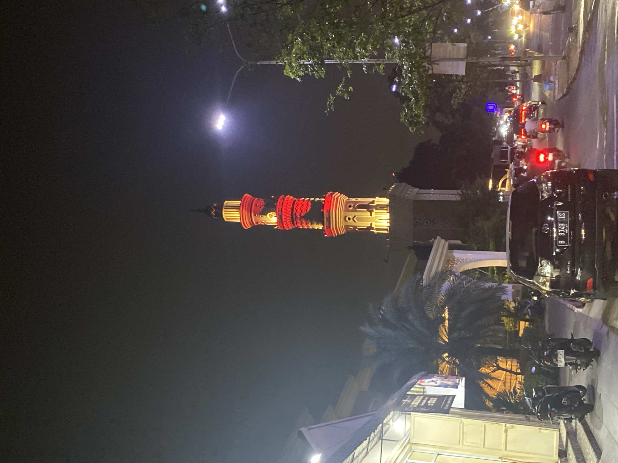 The image captures a street scene at night in Tangerang, Indonesia, with a prominent mosque minaret illuminated by red and yellow lights in the background. The minaret is tall and ornate, featuring multiple levels and decorative elements. In the foreground, a black SUV with a license plate ending in "LS" is parked on the right side of the road. To the left, a building with a white awning and large glass doors is visible, with two motorcycles parked in front. The street is moderately busy, with several other vehicles, including cars and motorcycles, driving by. A street lamp with bright white light is positioned to the right of the minaret, casting illumination on the surroundings. Trees with green foliage are present on the right side of the frame. The overall atmosphere suggests a vibrant urban environment at night.