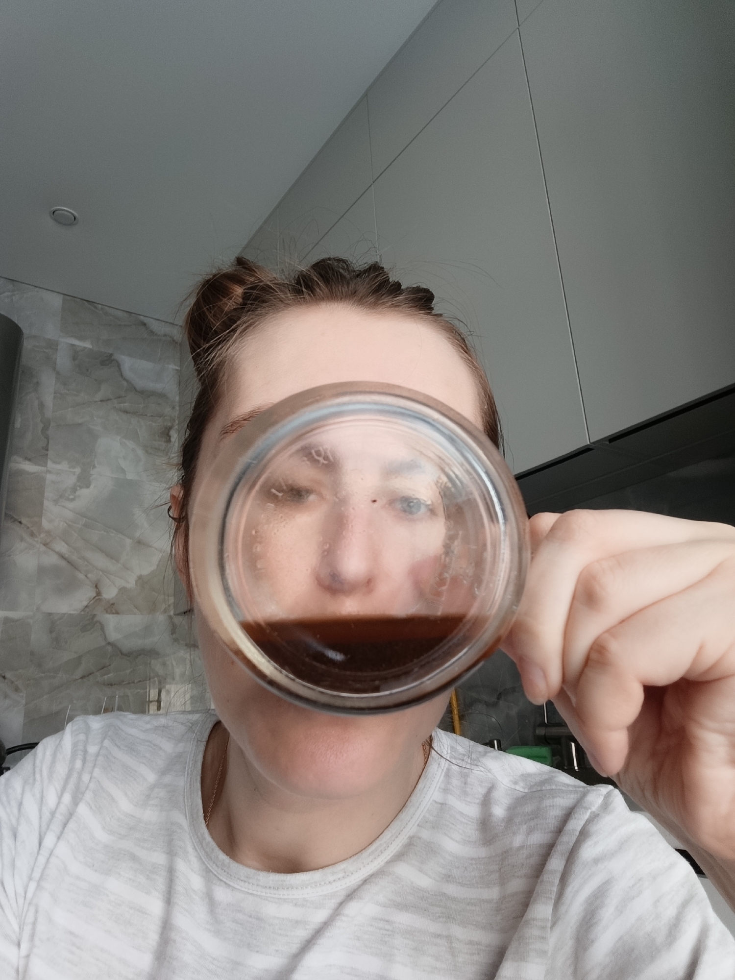 An individual with light skin and brown hair, tied back, is depicted in a close-up, high-angle image, consistent with a selfie, taken in an indoor setting within the city of Chaikovsky, Russia. The subject holds a clear glass containing a dark brown liquid, likely coffee or tea, to their face, with the glass partially obscuring the mouth and lower nose, indicating an action of drinking or preparing to drink. The subject's right hand is visible gripping the glass. They are wearing a light-colored t-shirt with subtle horizontal stripes and a thin, gold-colored chain necklace. A small black hoop earring is visible in the subject's left ear. The background features light-toned, large-format marble-patterned wall tiles on the left side, transitioning to smooth, light-grey paneled walls or cabinetry on the right. The ceiling above is light grey and includes a single recessed circular light fixture. Portions of what appear to be kitchen appliances, including a green-colored handle and metallic components, are discernible in the lower right background.