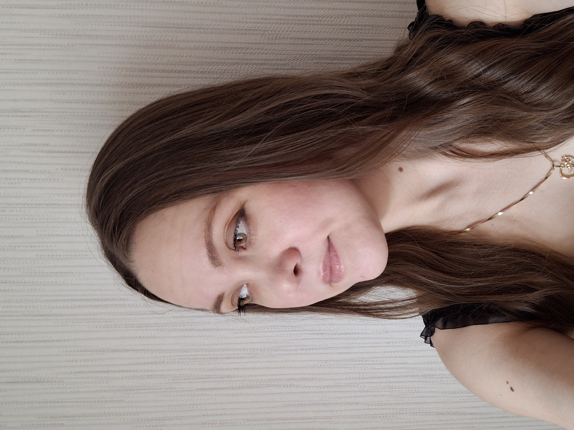 A young woman with long, light brown hair is visible from the chest up. She is wearing a black top with ruffled detailing. A thin gold necklace is around her neck. The woman is looking towards the viewer with her head slightly tilted. Her skin is fair, and she has light brown eyes and eyebrows. The background is a plain, light-colored wall with a subtle pattern. The location, Chaikovsky, Russia, cannot be directly confirmed from the image.