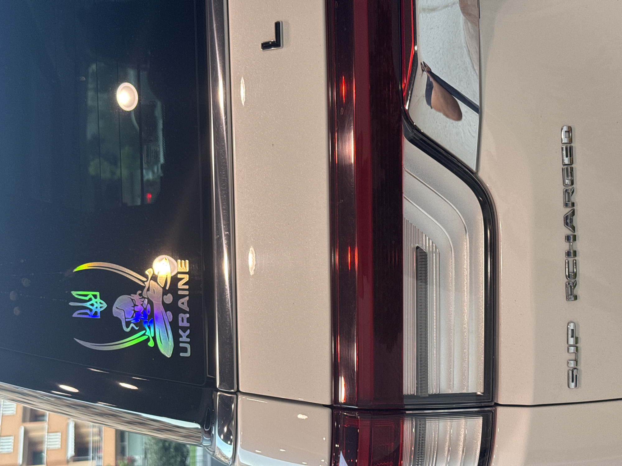A close-up, rear-view shot of a white vehicle's tailgate, featuring a holographic sticker on the rear window with the Ukrainian trident emblem and the word "UKRAINE" below. The sticker displays iridescent colors. The vehicle's taillights are a prominent red bar with chrome accents. Below the taillights, the chrome lettering "SUP RECHARGED" is visible. The background shows a blurry urban environment with buildings and greenery, suggesting it is located in Budva, Montenegro. The image has no people or discernible actions.