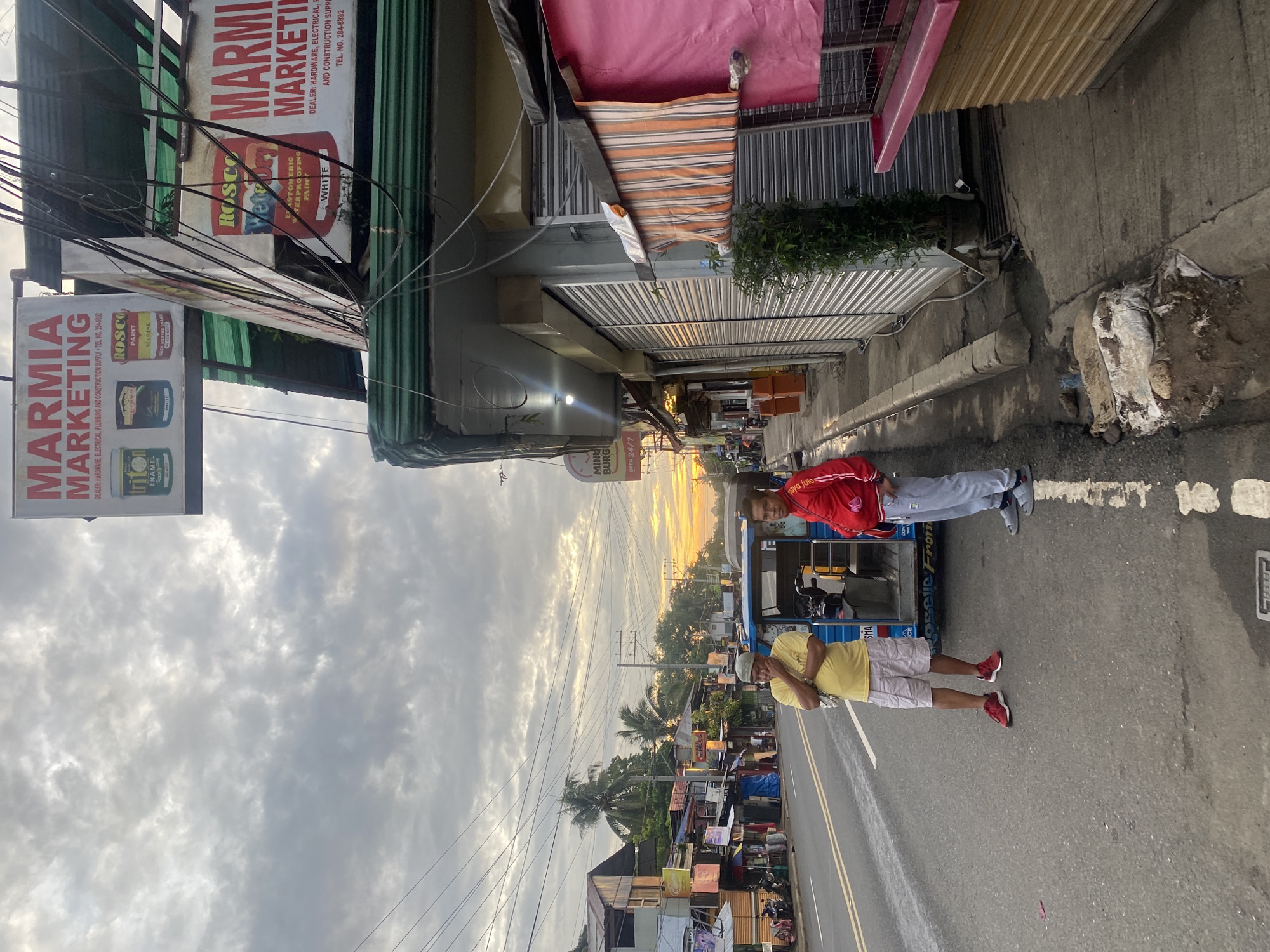 The image depicts a street scene in Davao City, Philippines, during what appears to be sunrise or sunset, given the warm, orange glow on the horizon and the overcast sky. The foreground is dominated by a paved road with a white lane marker. On the right side of the road is a sidewalk with a concrete curb. A small plant in a pot stands on the sidewalk. A pink canopy, striped orange and white awning, and a pink counter with a cage on it are visible along the storefronts.
Two men are present. One, wearing a yellow t-shirt and light-colored shorts, stands with his hand on his chin, looking towards the left of the frame. He is wearing red sneakers. Beside him, to his right, is a blue tricycle. The second man, wearing a red jacket and gray sweatpants, stands facing the viewer, looking slightly to his right. He is wearing gray sandals.
Behind the men, a blue tricycle is parked on the side of the road. Several storefronts line the left side of the street, many with roll-up metal doors that are closed. Signs for "MARMIA MARKETING" are visible on the buildings. These signs indicate that the business deals in hardware, electrical, plumbing, and construction supplies. The surrounding environment includes palm trees and residential or commercial structures, suggesting a mixed-use urban area. Power lines crisscross overhead. The overall impression is of a modest commercial street in a tropical climate.