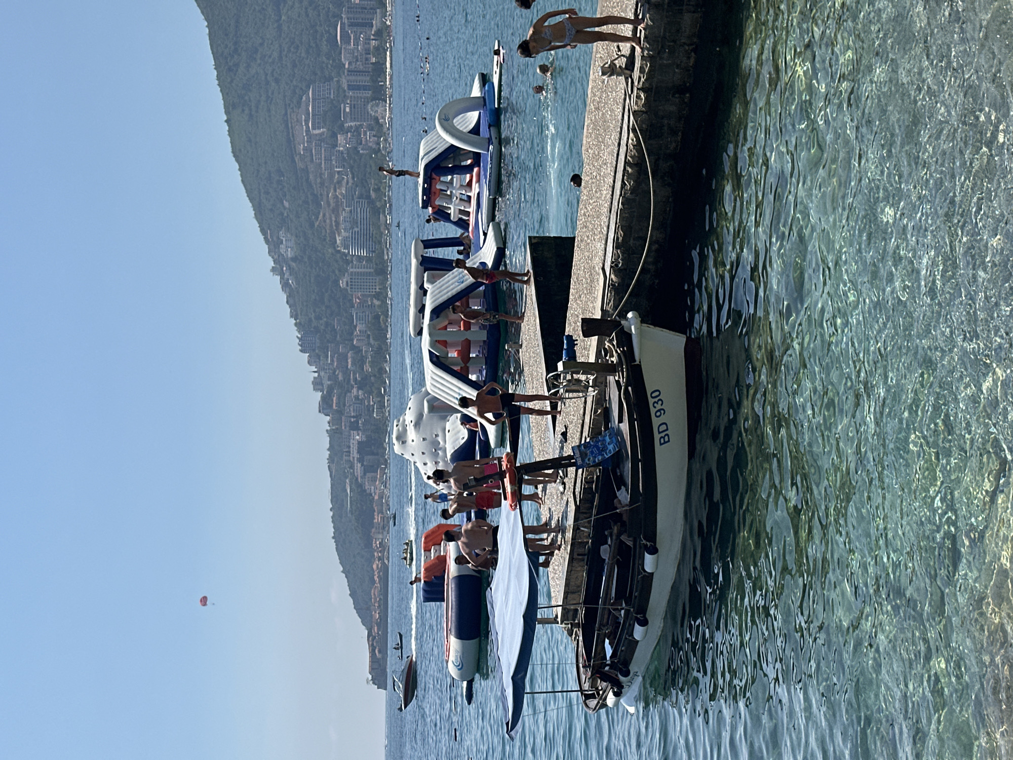The image depicts a busy scene at a beach in Budva, Montenegro, on a bright, sunny day. In the foreground, a small white boat with a blue canopy is docked at a stone pier. The boat has "BD 930" written on its side. Several people, mostly young men and boys, are standing on the pier near the boat, some in swim trunks, appearing to be waiting or socializing.
Further out in the turquoise water, a collection of inflatable water park structures is visible, including slides and climbing walls. Many individuals are interacting with these structures, with some children and adults swimming or playing in the water around them.
In the background, the cityscape of Budva unfolds, with buildings nestled on a lush green hillside that slopes down towards the sea. The hillside is dotted with more buildings, suggesting a densely populated coastal town. In the far distance, a boat is cruising on the water, and a red parasail is visible in the clear blue sky, indicating recreational activities. The overall atmosphere is one of summer leisure and fun.