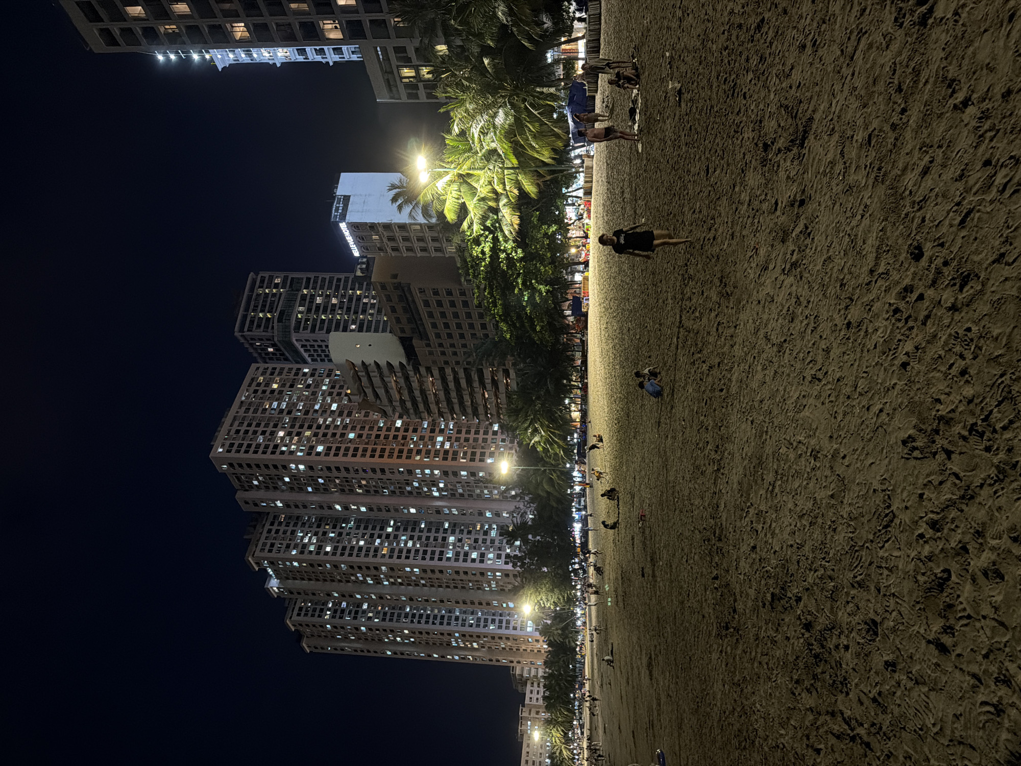 This is a nighttime photograph of a beach in Nha Trang, Vietnam, facing a row of tall apartment buildings. The beach is covered in sand with numerous footprints. Several people are visible on the beach, some walking, some sitting, and some appearing to be playing. One woman in a black shirt and shorts walks towards the camera. In the background, palm trees are illuminated by streetlights. Beyond the trees, the brightly lit windows of multiple high-rise apartment buildings create a stark contrast with the dark night sky. A sign on one of the buildings reads "BELLEVUE HOTEL." The scene captures a lively urban beach environment at night.