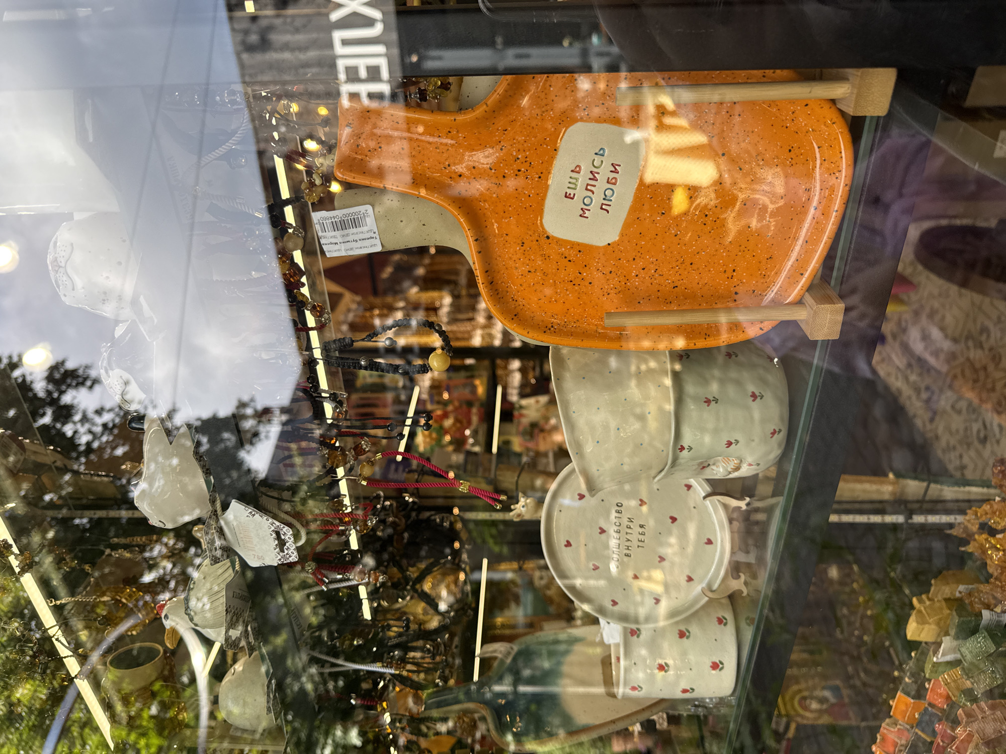 This is a medium-angle, eye-level, full-shot photograph taken through a glass window, likely a shop display, in Kaliningrad, Russia. The image is dominated by reflections of the sky and trees on the glass, creating a slightly hazy and layered effect.
In the foreground, on a glass shelf, are several ceramic items. To the left, a tall, slender vase with a teal and off-white gradient. Next to it, a white ceramic plate with red hearts and Cyrillic text in black, which reads "волшебство внутри тебя" (magic inside you). To the right of the plate are two white ceramic cups or bowls, one with red floral designs and the other with a similar red floral design and a small chicken illustration.
Further to the right, a bright orange ceramic platter with a speckled surface is propped up by two small wooden stands. On this platter, in a light-colored rectangular inset, are Cyrillic words in colored lettering: "ЕШЬ МОЛИСЬ ЛЮБИ" (Eat Pray Love). Behind these ceramic items, various beaded necklaces and other trinkets are displayed, hanging from above.
In the background, visible through the glass and reflections, appear to be more shelves filled with various objects, including what look like small decorative ceramic birds and other small gift items. The overall impression is that of a whimsical souvenir or gift shop.