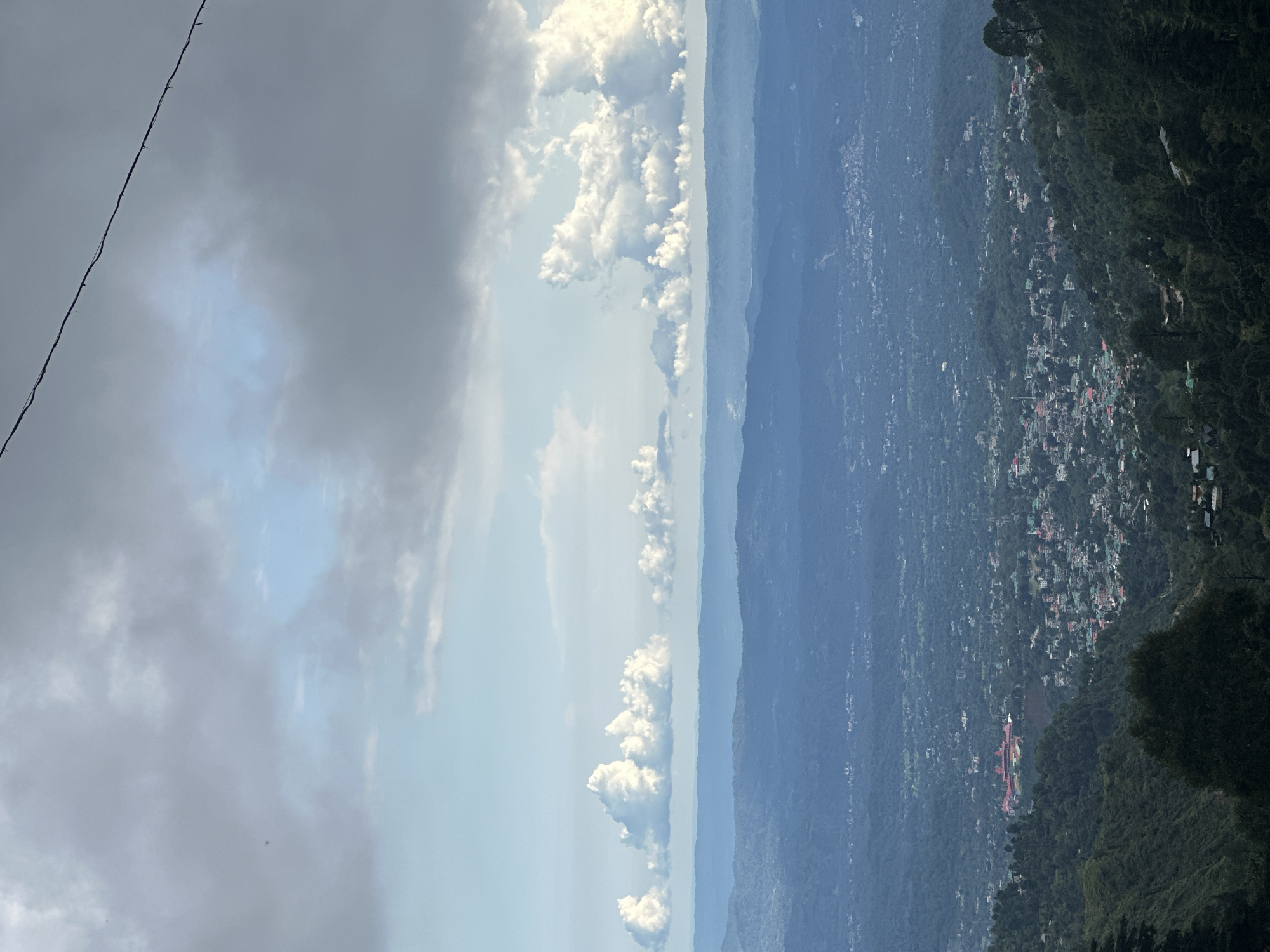 The image depicts a wide aerial view of the city of Chouhla, India, nestled amidst rolling, forested hills under a partly cloudy sky. The foreground is dominated by dark green, dense foliage and trees on a steep slope, with hints of structures visible at the very bottom edge.
In the mid-ground, the city of Chouhla spreads out across the landscape. Numerous buildings with red roofs are clustered together, interspersed with patches of greenery. A prominent, large, red-roofed complex, likely a significant institution or landmark, is visible on a hillside overlooking the city. The city itself appears to extend into the distance, becoming increasingly hazy and bluish due to atmospheric perspective.
The background consists of several layers of receding hills, each appearing fainter and more blue than the last, creating a sense of depth and vastness. A distinct line of fluffy white clouds hovers above the furthest hills, with darker, more diffuse grey clouds filling the upper portion of the sky. A thin, dark wire runs diagonally across the upper right portion of the image, suggesting the presence of overhead power lines or a similar man-made obstruction. There are no visible people or animals in the frame. The overall impression is one of a sprawling urban settlement integrated into a lush, mountainous natural environment.