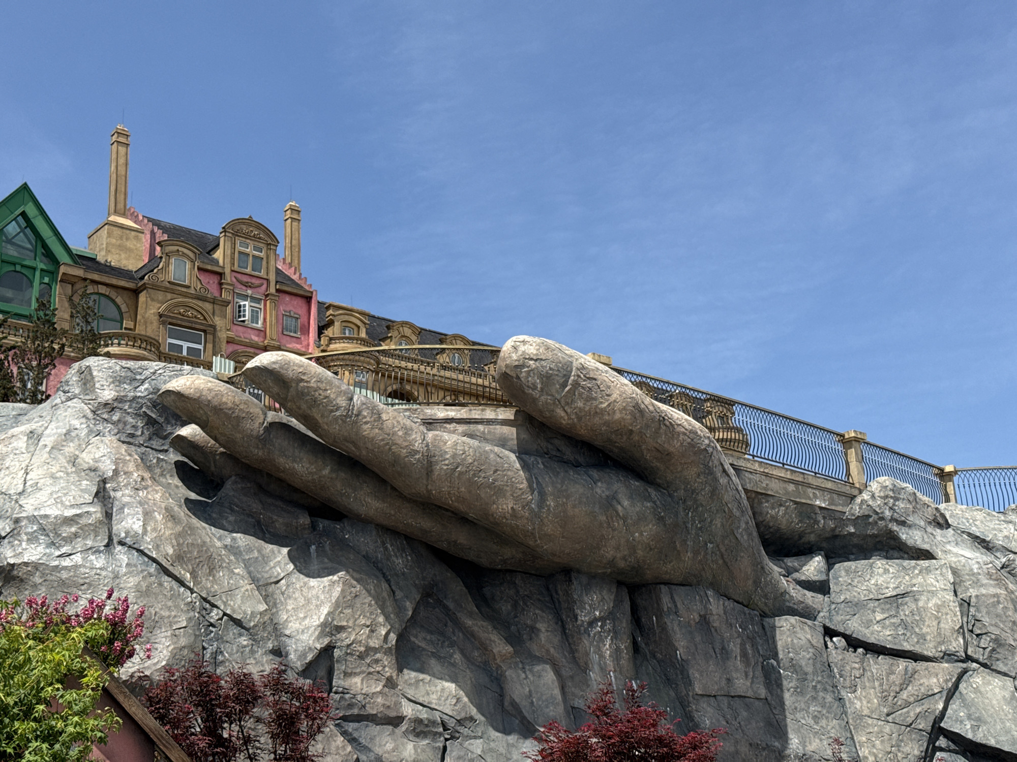 The image shows a large stone sculpture of a hand emerging from a rocky cliff face, located in the city of Đà Nẵng, Vietnam. The hand supports a pedestrian walkway with a decorative metal railing. Behind the sculpture, a multi-story building with various architectural styles, including a green-roofed section and pink facade, is visible against a clear blue sky. Some vegetation is visible at the base of the rocky formation.