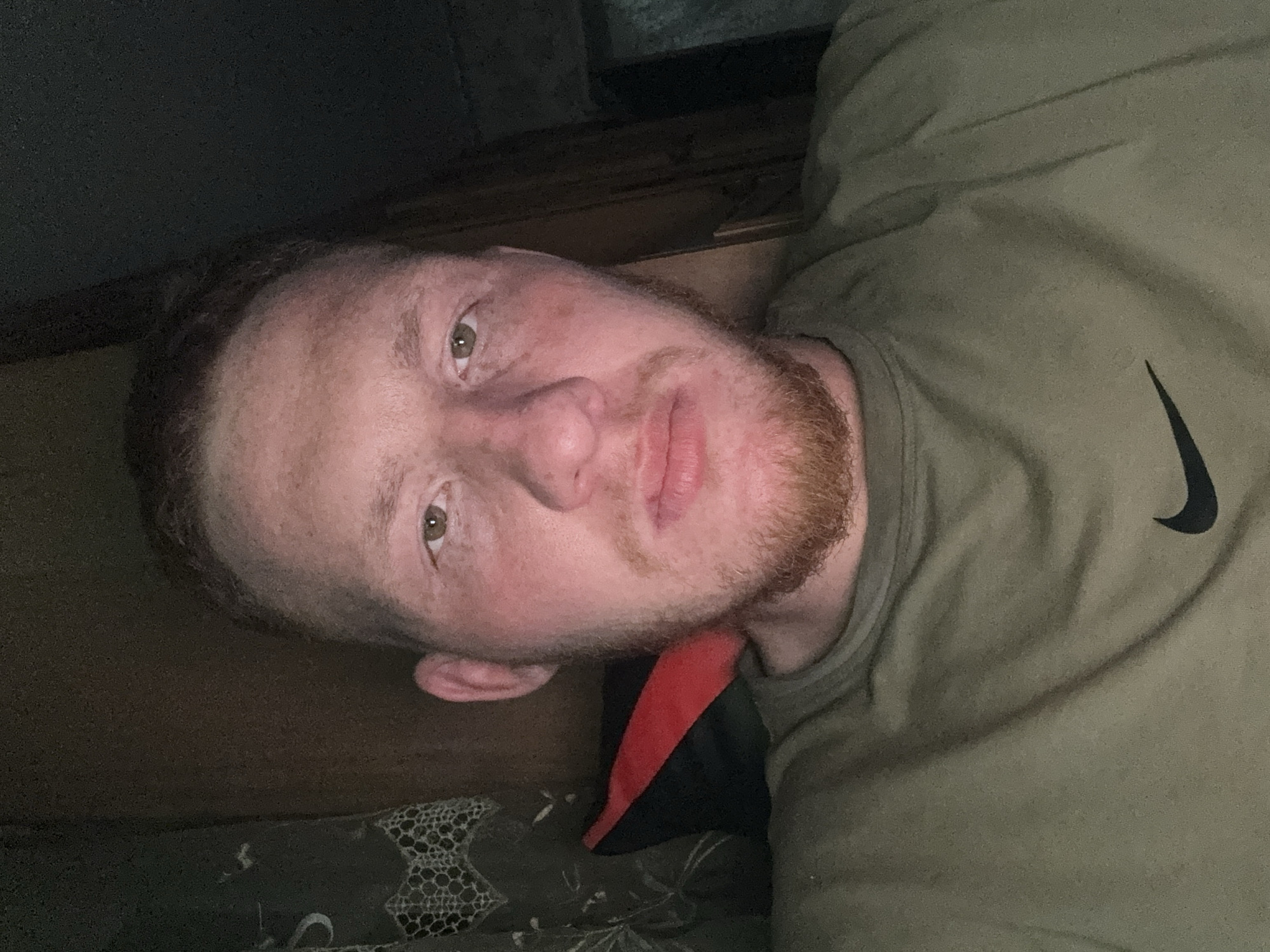 A close-up selfie shows a man with fair skin, light brown hair, and a reddish-brown beard. He is wearing an olive green t-shirt with a black Nike swoosh logo on the right side of the chest. The background is dark and indistinct, revealing a wooden surface behind his head. A black and red object is visible near his neck. The man is indoors, lying down. The scene is undetailed, and there is no indication of Makiivka, Ukraine.