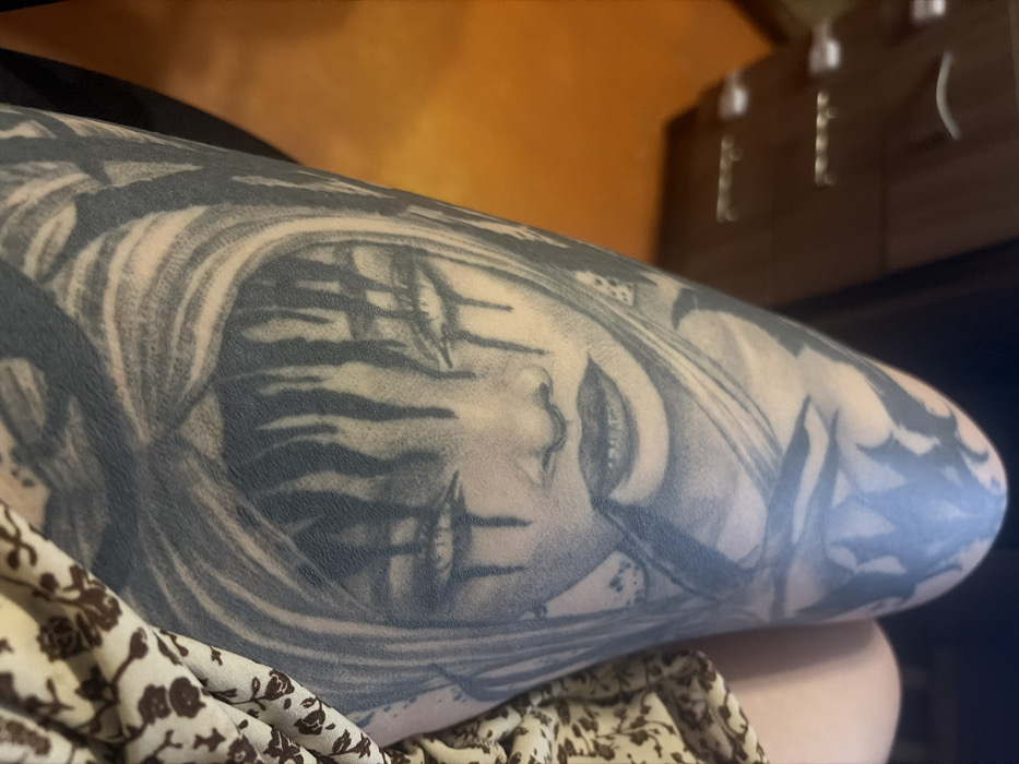 The image depicts a close-up view of a person's upper leg, likely a thigh, featuring an extensive grayscale tattoo, in what appears to be an indoor setting in Chaikovsky, Russia. The tattoo occupies the majority of the visible leg area and portrays an inverted stylized face with prominent dark eye markings or tears extending downwards, defined lips, and surrounding elements resembling drapery or hair. Above the face, closer to the knee, other abstract or organic tattoo patterns are visible. To the right of the leg, a portion of a light-colored garment with a small, repeating dark brown floral or abstract pattern is visible, suggesting the subject is wearing a dress, skirt, or patterned shorts. In the blurred background to the upper left, a brown wooden chest of drawers with three visible drawers and silver-colored handles is present. Below the furniture, a section of an orange-colored floor is visible. The scene is lit, with a slight sheen on the tattooed skin near the knee. The image is captured from a slightly elevated, downward-angled perspective.