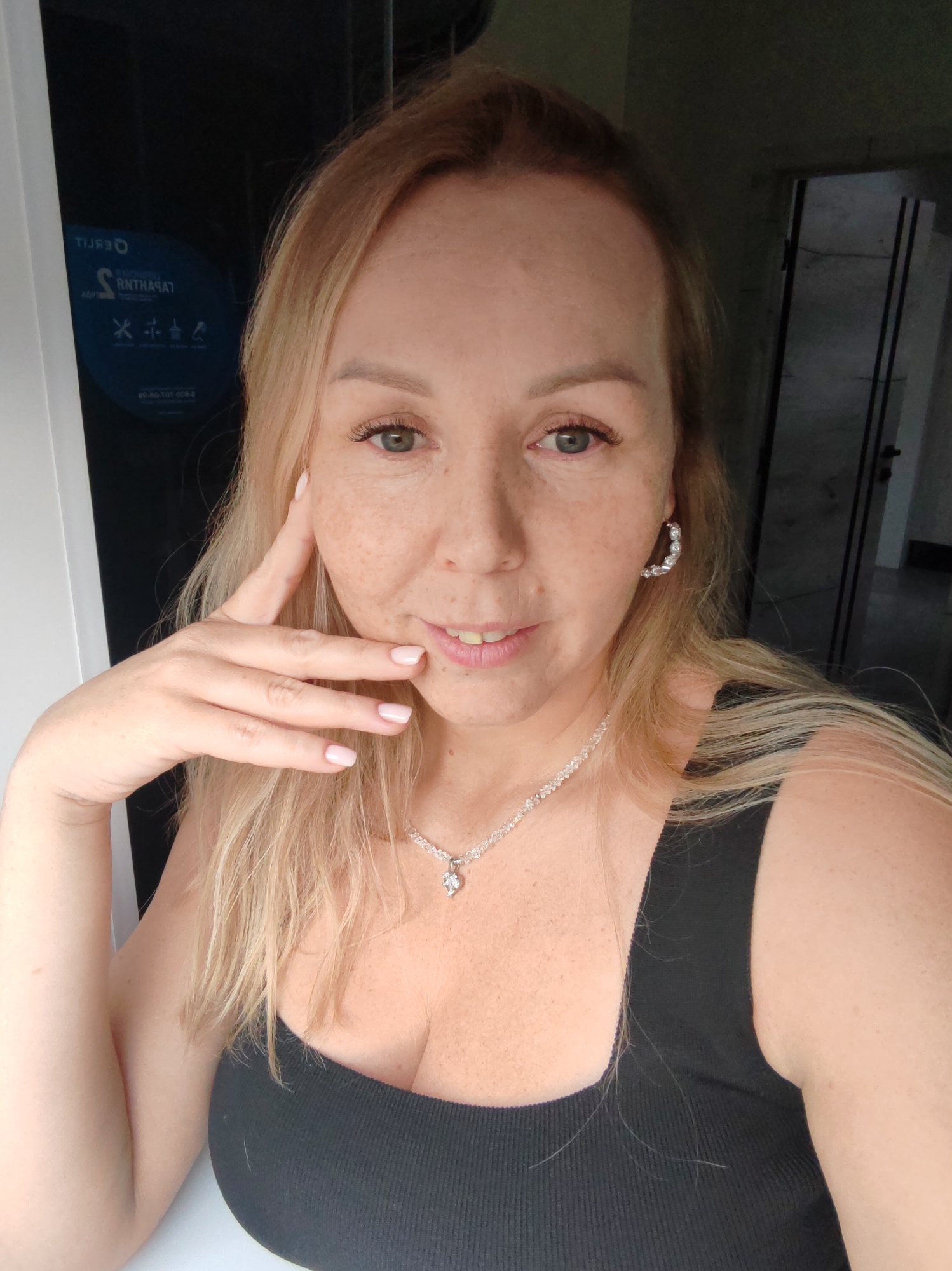 A selfie of a woman with blonde hair, freckles, and light-colored eyes. She's wearing a black tank top, a diamond necklace, and hoop earrings. Her left hand is touching her face near her cheek. Behind her is a partially visible doorframe leading to what appears to be a room with a dark interior. To the left, a black panel has what seems to be a sticker or sign with text in Russian. Based on the text "Тамара", it can be inferred that the location is Chaikovsky, Russia.