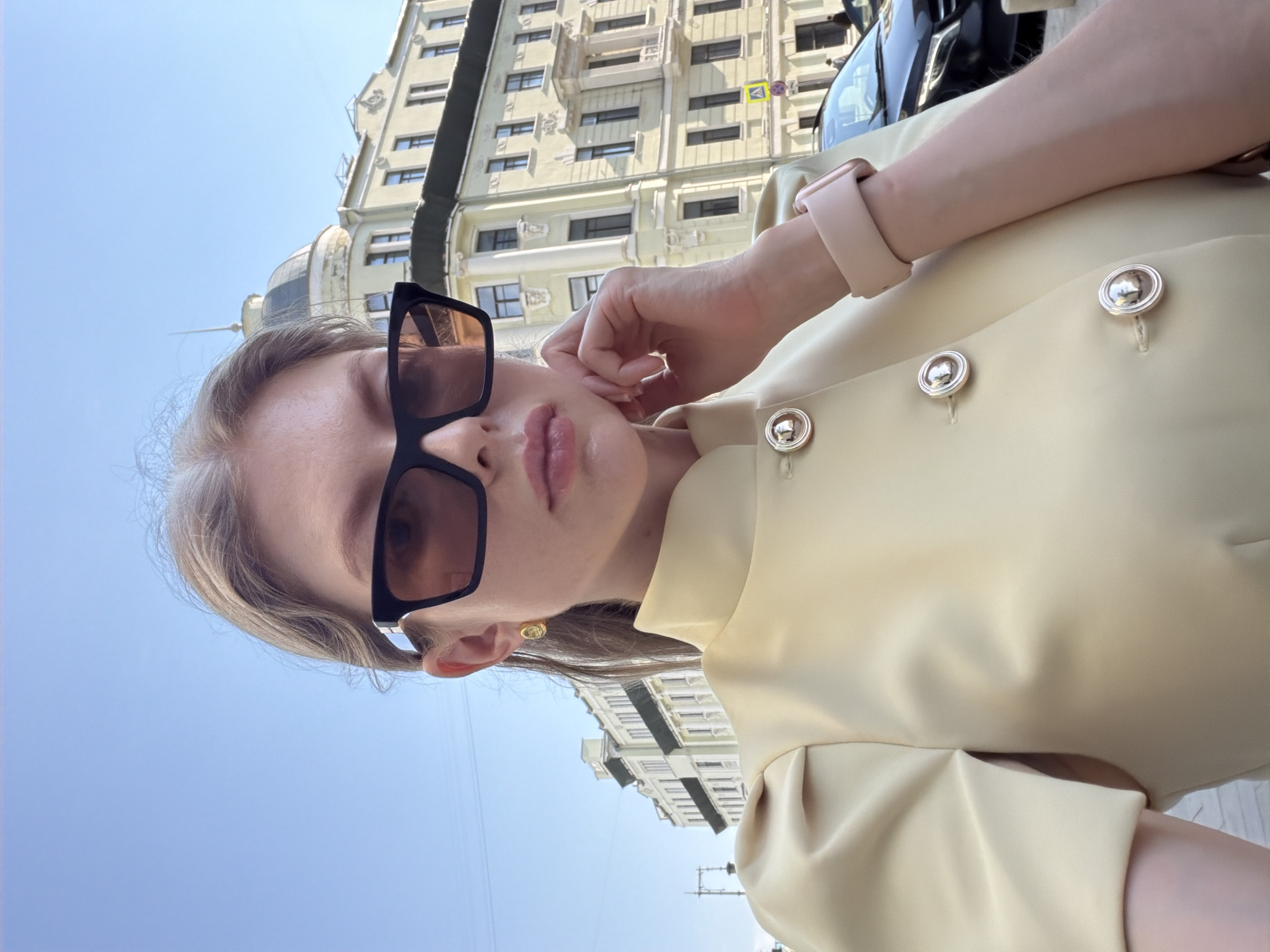 A pale-skinned woman with light brown hair stands outside in front of a building in Moscow, Russia, looking down at the camera. She is wearing oversized black sunglasses and a beige-colored top with silver buttons. A light pink smartwatch is visible on her left wrist. The building behind her is multi-storied and features a beige exterior with symmetrical windows and architectural details. Above her head, a clear blue sky is visible. A black car or SUV is partially visible in the upper right corner. The scene appears to be taken during the daytime, with bright, natural lighting.