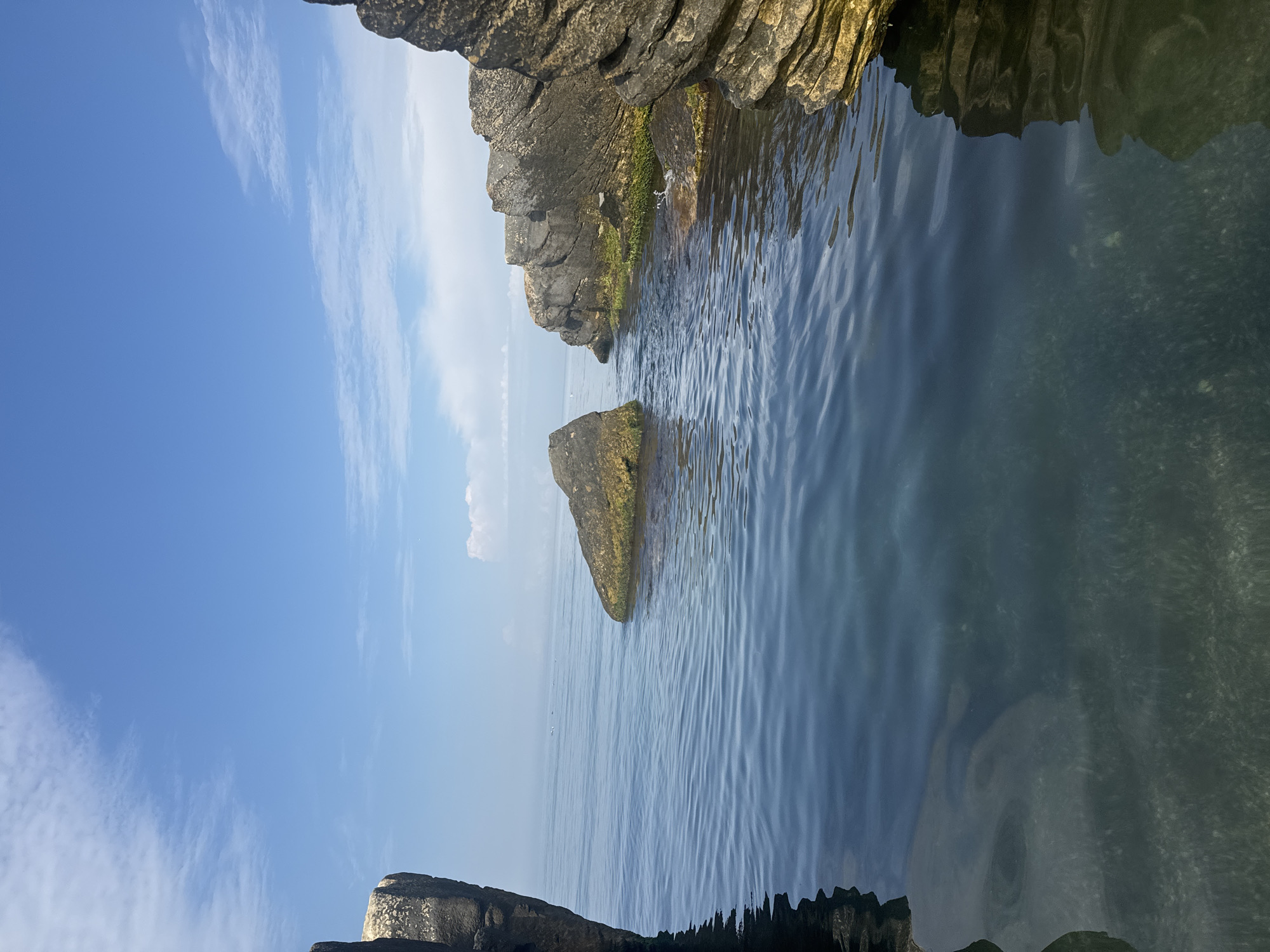 The image captures a serene coastal scene with clear blue water and a bright sky. In the foreground, the water is calm, reflecting the sky and the rocky shoreline. Several large rocks, some covered in green moss, jut out of the water and line the coast. In the distance, a faint haze obscures the horizon, with a few small white clouds scattered across the sky. A group of small white birds are visible on the water in the middle distance. The overall impression is one of natural tranquility. This scene is characteristic of the coastline near the city of Aktau, Kazakhstan, known for its unique rock formations and the Caspian Sea.