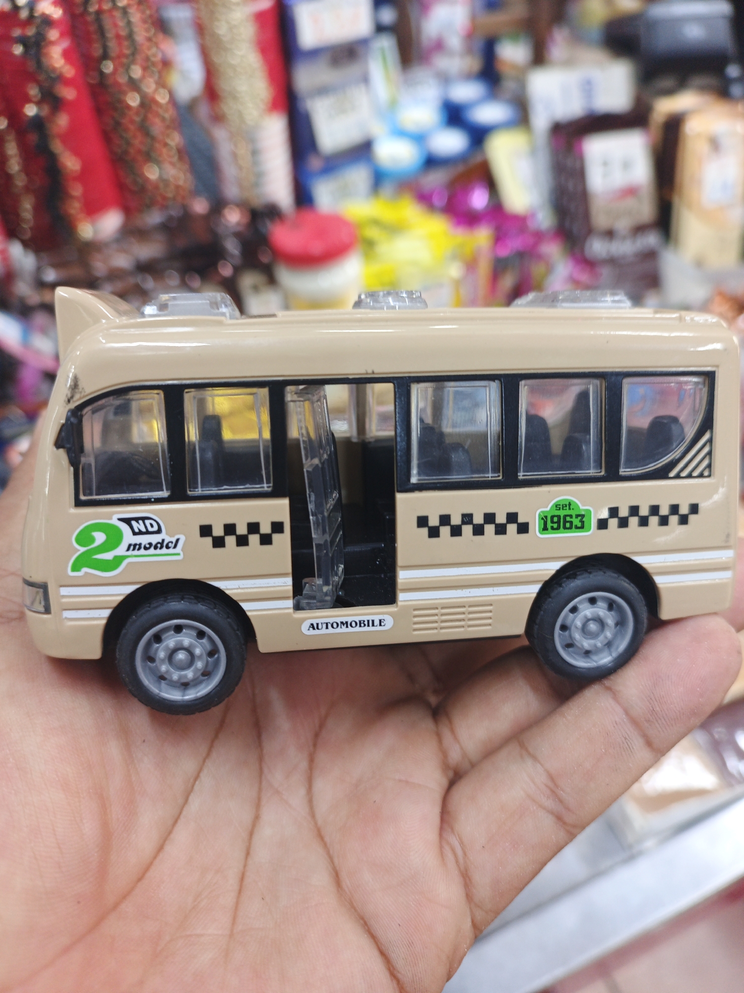 A close-up shot features a light tan-colored toy bus held in a person's hand. The toy bus has black-framed windows, with the front passenger door ajar, revealing black seats inside. It is equipped with gray-rimmed wheels and black tires.
Distinctive markings on the side of the bus include a large green "2 ND model" logo near the front, and a black and white checkered pattern running along the lower side. Towards the rear, a green oval displays the text "set. 1963". A small label reading "AUTOMOBILE" is visible below the checkered pattern, near the front wheel. The hand holding the toy shows visible palm lines.
The background is blurred, indicating an indoor retail environment, likely a shop or market stall in Tinaquillo, Venezuela. Various colorful items are vaguely discernible, such as red and gold beaded items on the left and packaged goods, possibly snacks or toiletries, on the right. The scene is illuminated by artificial indoor lighting. No specific activity beyond holding the toy is taking place, and no emotions are discernible. The time of day cannot be determined.