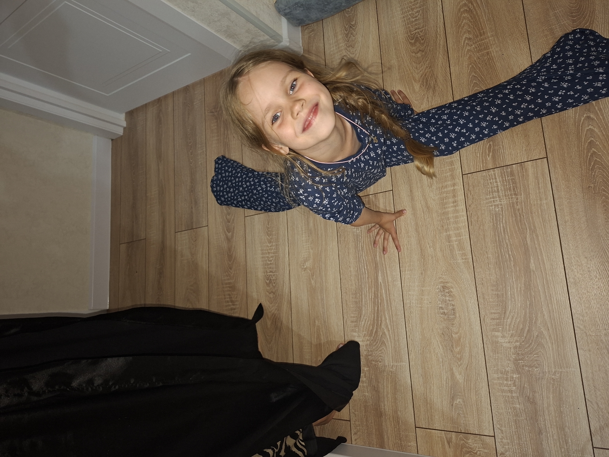 A young girl with blonde hair is lying on a wooden floor, looking up at the camera with a smile. She is wearing a blue long-sleeved shirt with a white floral pattern and dark blue pants with the same pattern. Her arms are outstretched, and one hand is near the camera.
The room has light-colored wooden floorboards and a white baseboard along the wall. In the background, there is a doorway with a white door. Part of a black garment is visible in the foreground, obscuring the lower portion of the image.
The lighting suggests it is daytime, and the setting appears to be indoors. The girl's expression conveys happiness and playfulness. The location context is Izhevsk, Russia. There is no visible text in the image.