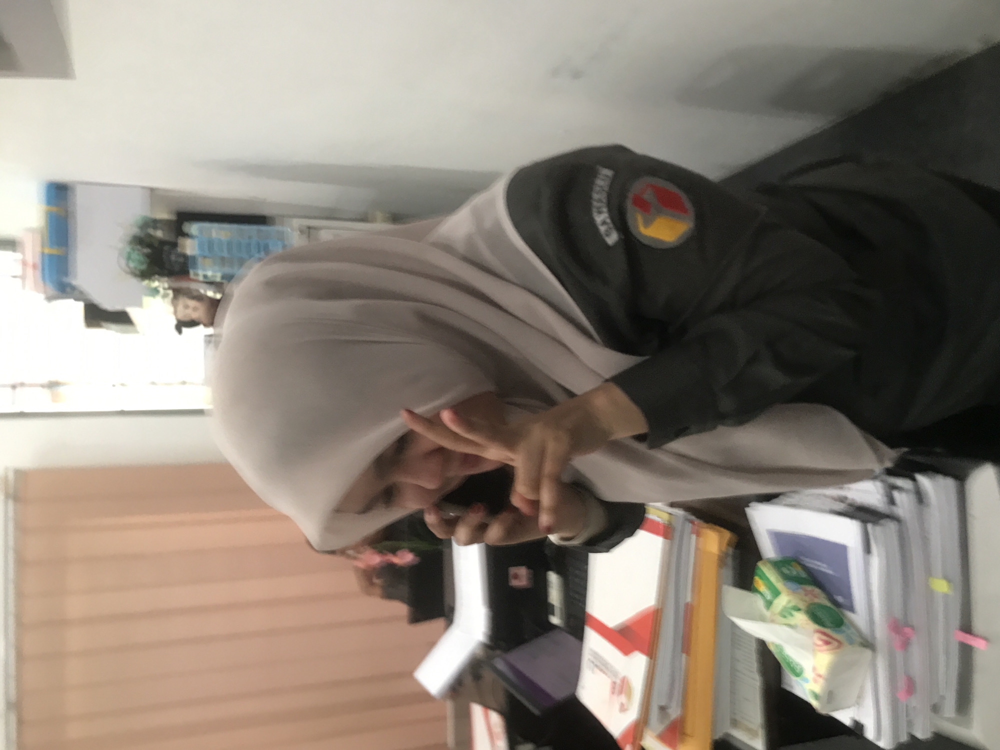 A woman wearing a light-colored headscarf and a dark uniform with a circular patch on the sleeve is holding a cell phone to her face. She is sitting at a desk with several stacks of papers, folders, and a box of tissues. The scene is indoors. It is not possible to determine the location in the city of Lhokseumawe, Indonesia, from the image.