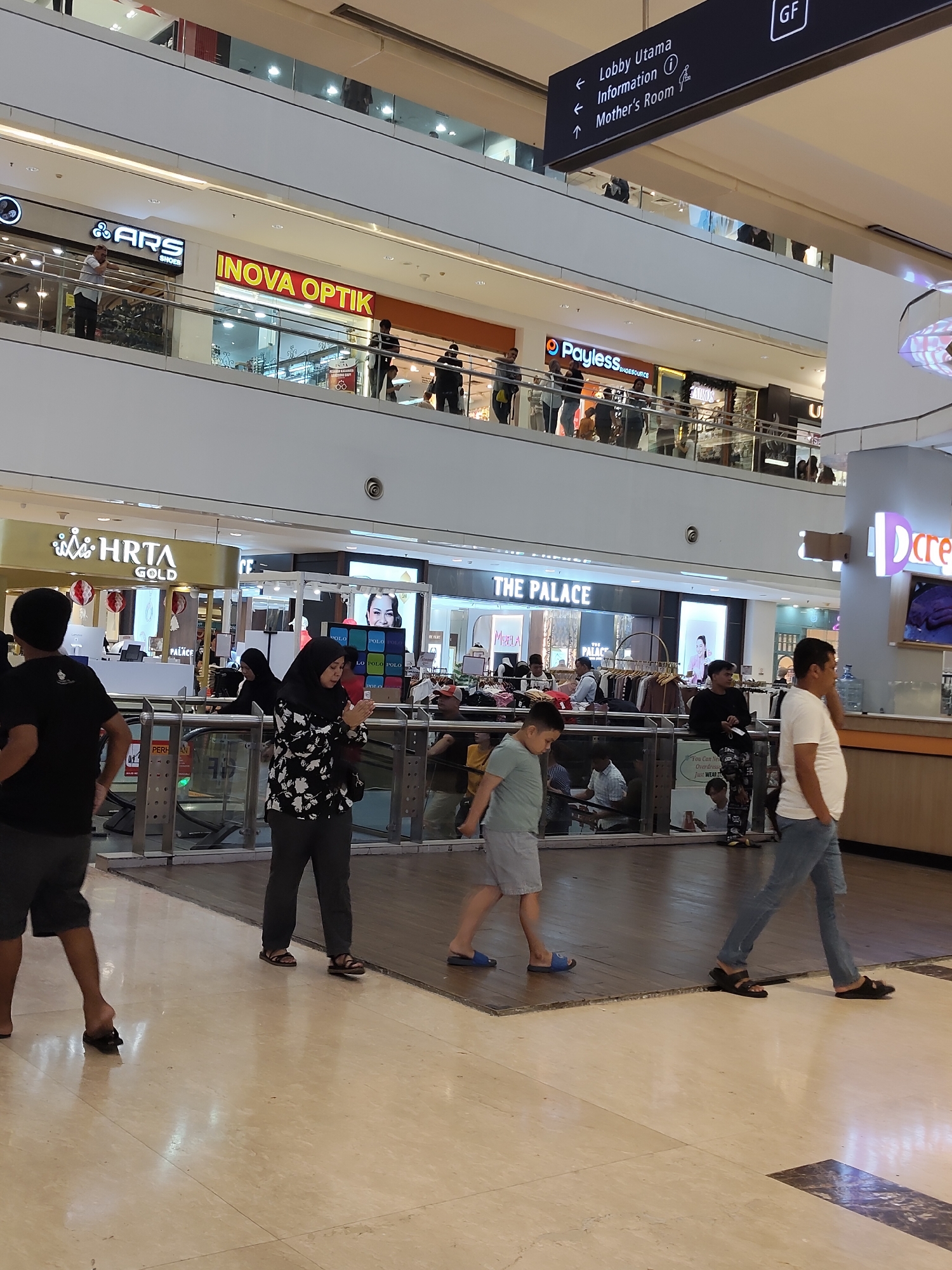 The image depicts the interior of a multi-story shopping mall in the city of Medan, Indonesia. On the upper levels, signage indicates stores such as "ARS SHOES" and "INOVA OPTIK." A prominent directional sign points towards "Lobby Utama," "Information," and "Mother's Room," with "GF" (Ground Floor) also indicated.
On a lower level, more store fronts are visible, including "HRTA GOLD" and "THE PALACE." Several individuals are present within the mall. In the foreground, a person in a black t-shirt and shorts walks away from the camera. Further to the right, a person wearing a white t-shirt and blue jeans walks towards the right, talking on a mobile phone. Between them, a child in a light blue t-shirt and shorts walks towards the left, and a woman in a black and white patterned shirt and dark pants, wearing a hijab, stands facing the camera. Other shoppers are seen on the levels above and in the background. The mall environment features polished floors, glass railings, escalators, and various retail displays.