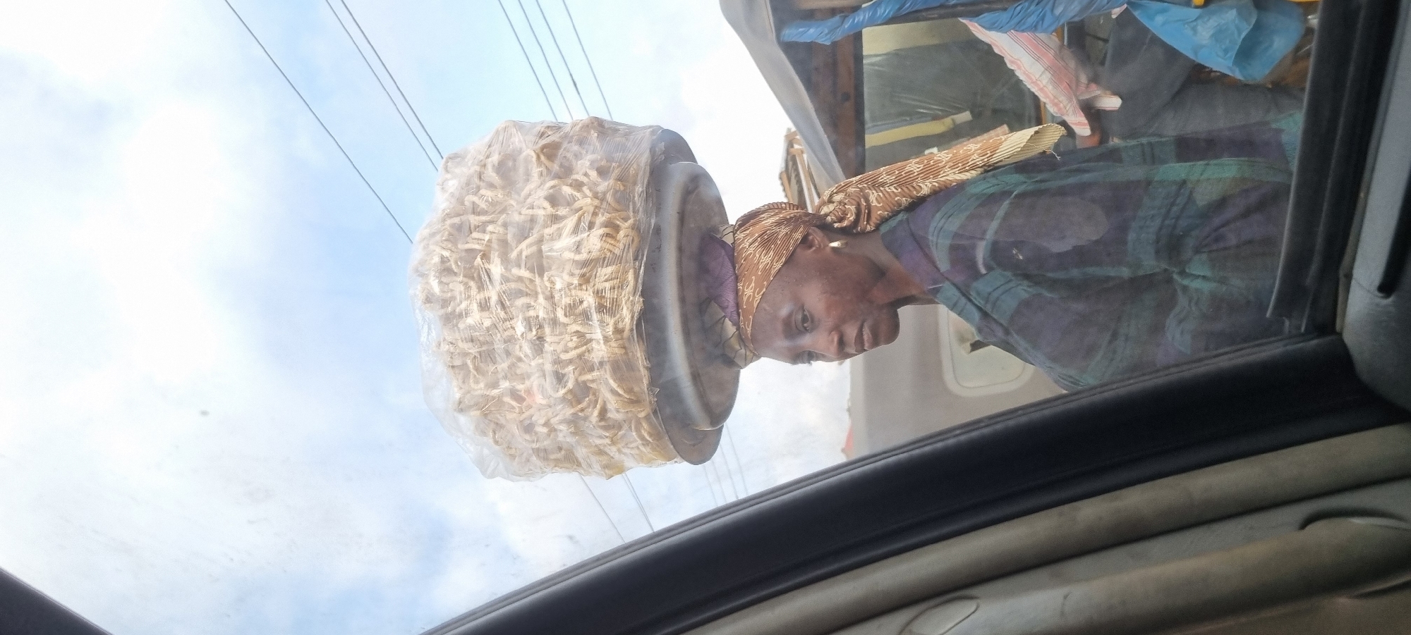 A woman is shown from the chest up, carrying a large, clear plastic-wrapped load of what appears to be dried fish on her head. She is wearing a patterned headscarf and a plaid shirt. She is looking to her right, slightly downwards. The scene is viewed from inside a vehicle, with part of the car's interior visible in the foreground on the left. Behind her, other vehicles and people are visible in what appears to be a busy street environment. This image was taken in Otta, Nigeria.