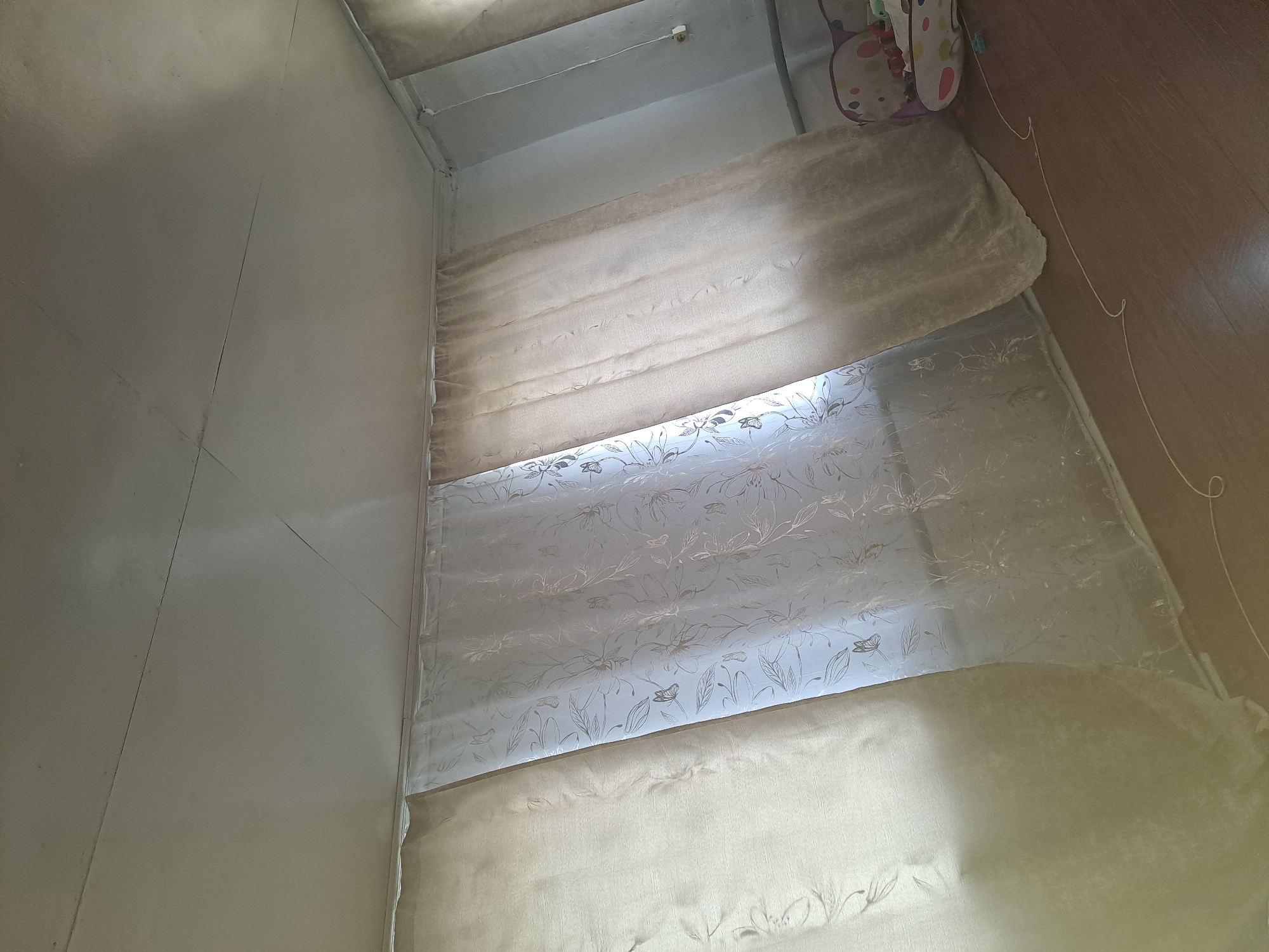 The image shows a room with curtains covering a window. The curtains are of two types: a sheer white curtain with a floral and butterfly pattern, and a thicker, opaque beige curtain. The ceiling appears to be made of drywall and has visible seams and cracks. The floor is made of wood. In the background, a small part of a child's seat with colorful patterns is visible. There are no people visible in the image. The location is a room in the city of Стрельцы, Russia.