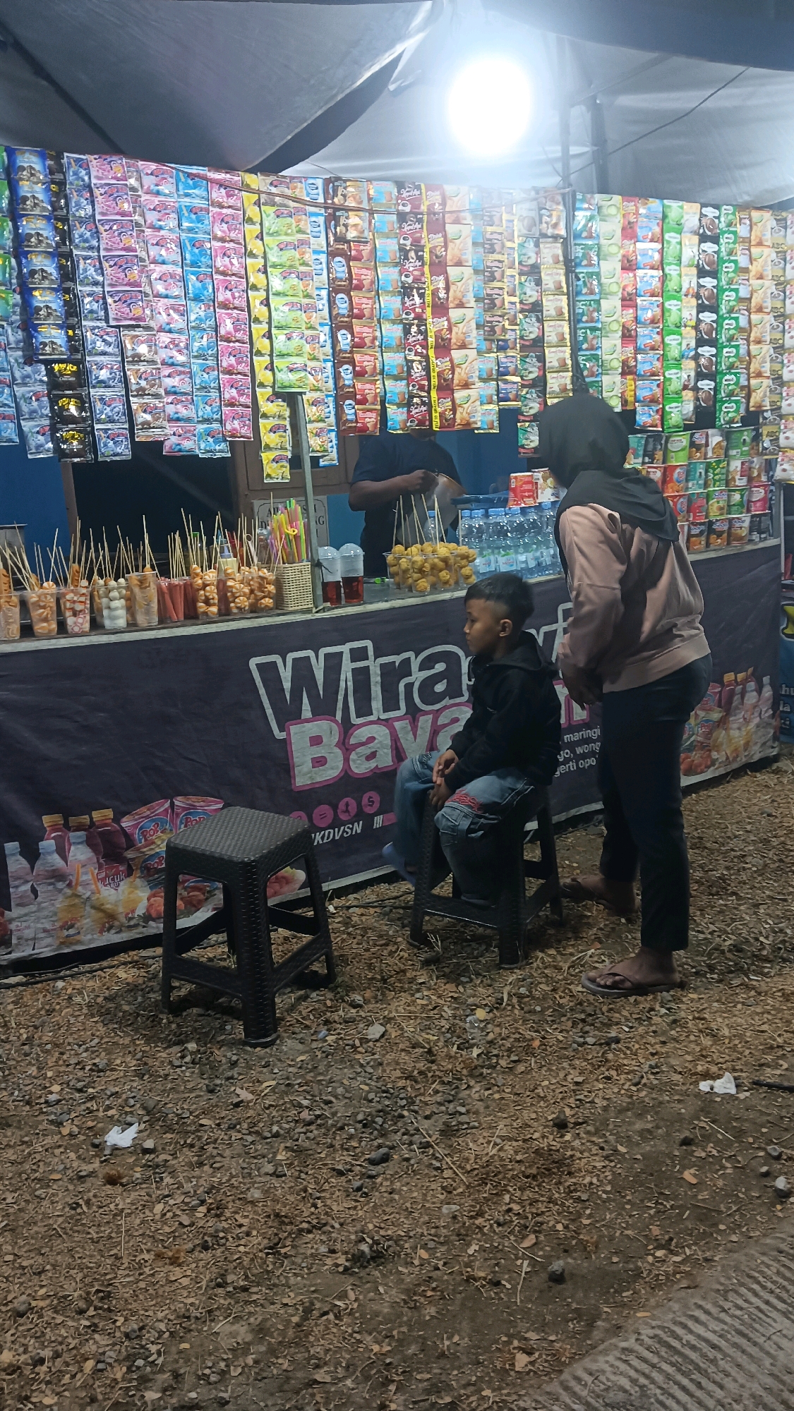 A young boy sits on a black stool in front of a food stall in Newung, Indonesia. The stall is decorated with a banner that reads "Wira Baya." Behind the boy, a woman in a hijab and a person in a black shirt stand at the counter of the stall, where various snacks and drinks are displayed. The stall is illuminated by a bright light from above. The ground is covered in gravel and fallen leaves.