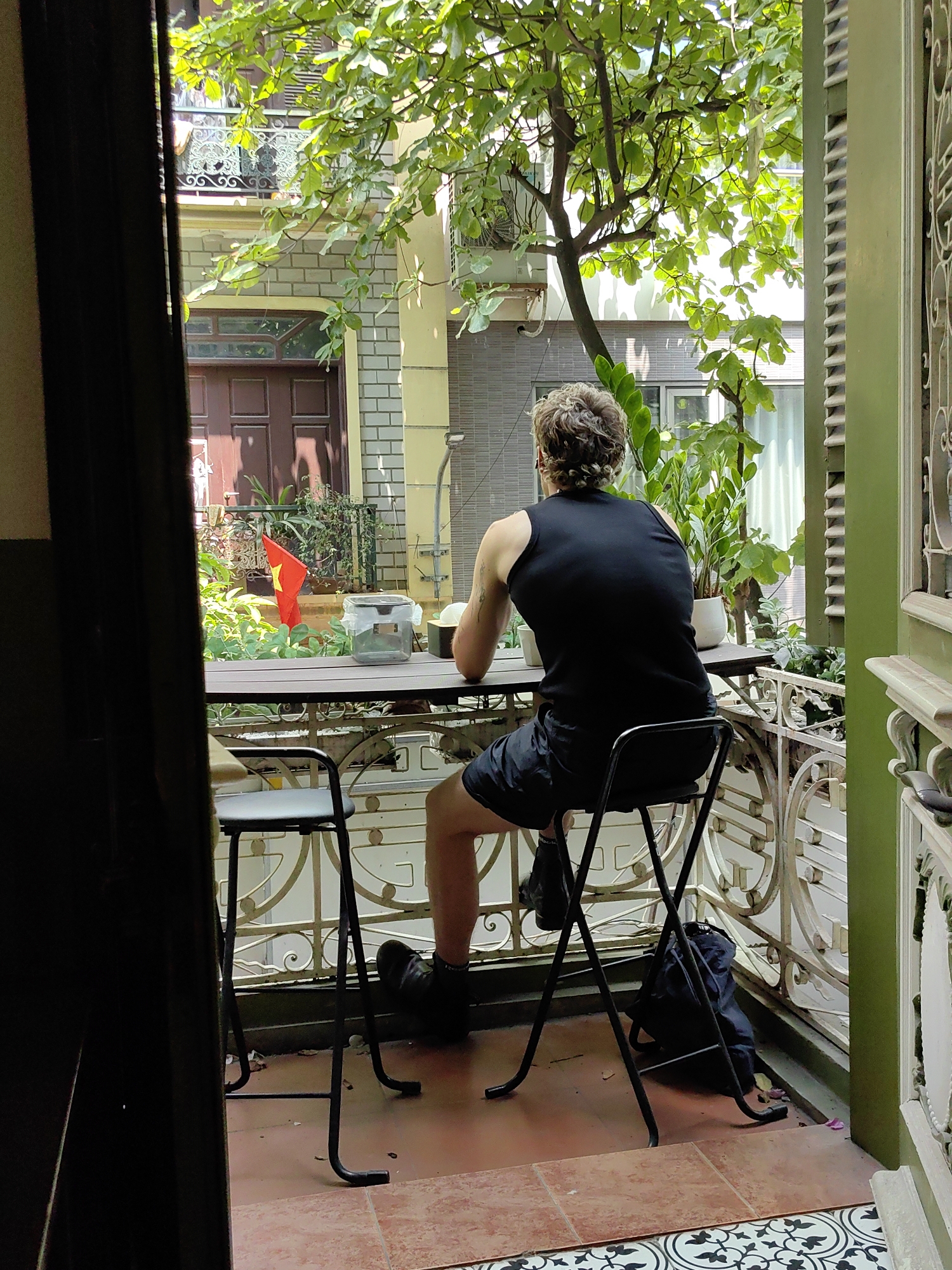 A man with curly, light brown hair, wearing a black sleeveless top and black shorts, is seated on a black metal stool at a tall, dark wooden table on an outdoor balcony. He is facing away from the camera, looking out towards the view. A white cup sits on the table in front of him, and a black backpack rests on the tiled floor beneath his stool.
The balcony features ornate white metal railings with intricate patterns. The floor is tiled in terracotta tones, with a decorative black and white patterned tile section closer to the foreground. Lush green foliage from trees and potted plants surrounds the balcony, providing a sense of greenery and partial shade. Through the dense leaves, a small red Vietnamese flag with a yellow star is visible.
In the background, parts of buildings with windows and a brown wooden door are visible, featuring light-colored brickwork and rendered sections. Bright natural sunlight illuminates the scene, creating strong highlights and shadows, suggesting a clear day, likely mid-morning or afternoon. The setting evokes a peaceful outdoor space, possibly a cafe or a home balcony, in Hà Nội, Vietnam. There is no visible text in the image.