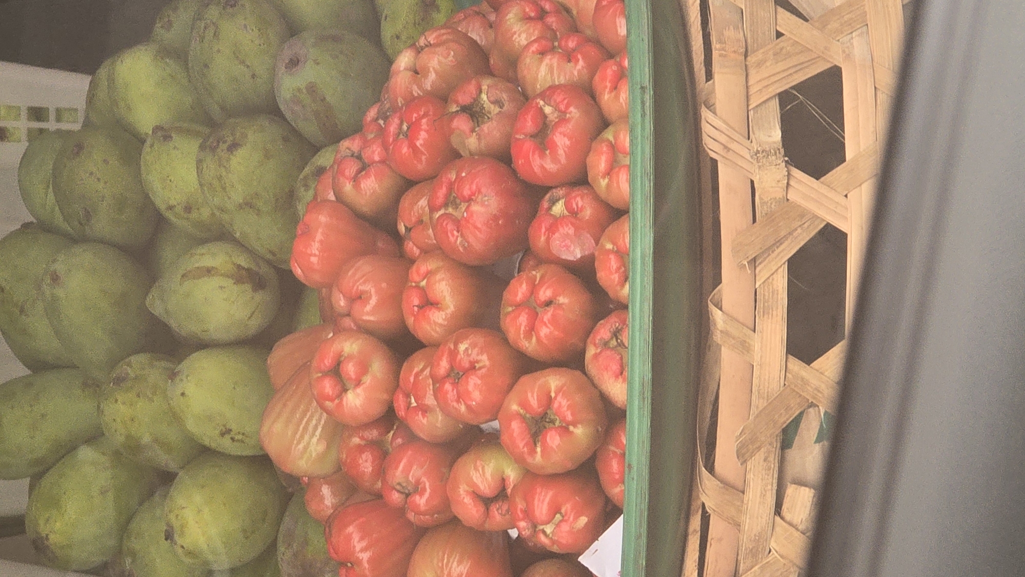 This image displays a vibrant assortment of fresh tropical fruits, likely from a market in Ubud District, Indonesia. The scene is dominated by two large piles of different fruits, arranged side-by-side.
On the left, there is an extensive mound of green, oval-shaped fruits, which appear to be unripe mangoes or a similar tropical variety. These fruits exhibit some natural blemishes and variations in their smooth, green skin.
To the right, a contrasting and equally abundant stack of bright red, bell-shaped fruits is prominently featured. These are clearly identifiable as rose apples (also known as water apples or jambu air), characterized by their waxy, deep red skin and distinct shape. A thin, dark green divider or container edge separates the green fruits from the red ones.
Further to the right, a section of a traditional, light brown woven bamboo basket is visible, showcasing intricate patterns created by the interwoven strips. This element adds a cultural touch, suggesting local craftsmanship. The far right edge of the frame is partially obscured by an opaque, dark grey or black object, possibly a curtain or part of a market stall structure.
The overall lighting is bright and natural, indicating it is daytime. The fruits are neatly stacked, suggesting they are on display for sale. No people or specific activities are visible, and there is no discernible text in the image.