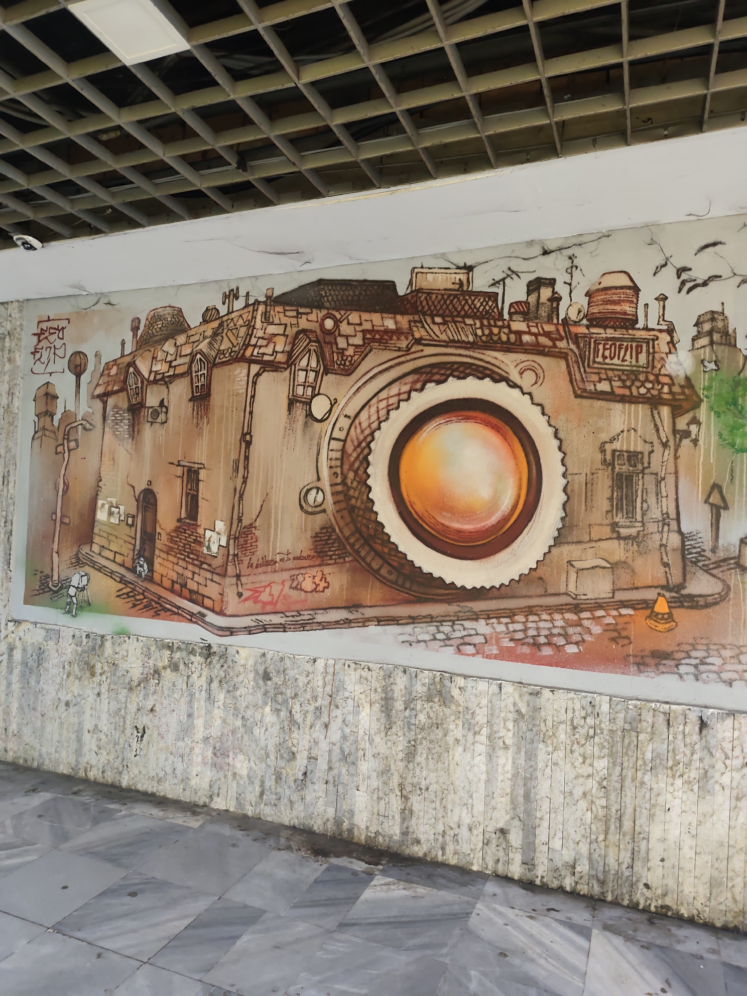 The image shows a mural on a textured, stone-like wall under a grid-patterned ceiling, possibly inside a building or covered walkway. The mural depicts a large, stylized camera formed from buildings and urban elements. The camera's lens is a prominent orange and white circle, resembling an eye. The buildings have windows, doors, roofs, and chimneys, with some featuring graffiti. At the bottom of the mural, there is a painted streetscape with a figure holding a camera and another figure walking down the street. To the right of the camera building is a painting of a cone. The flooring in front of the wall is a gray marble tile pattern. The location is Unknown, Unknown.