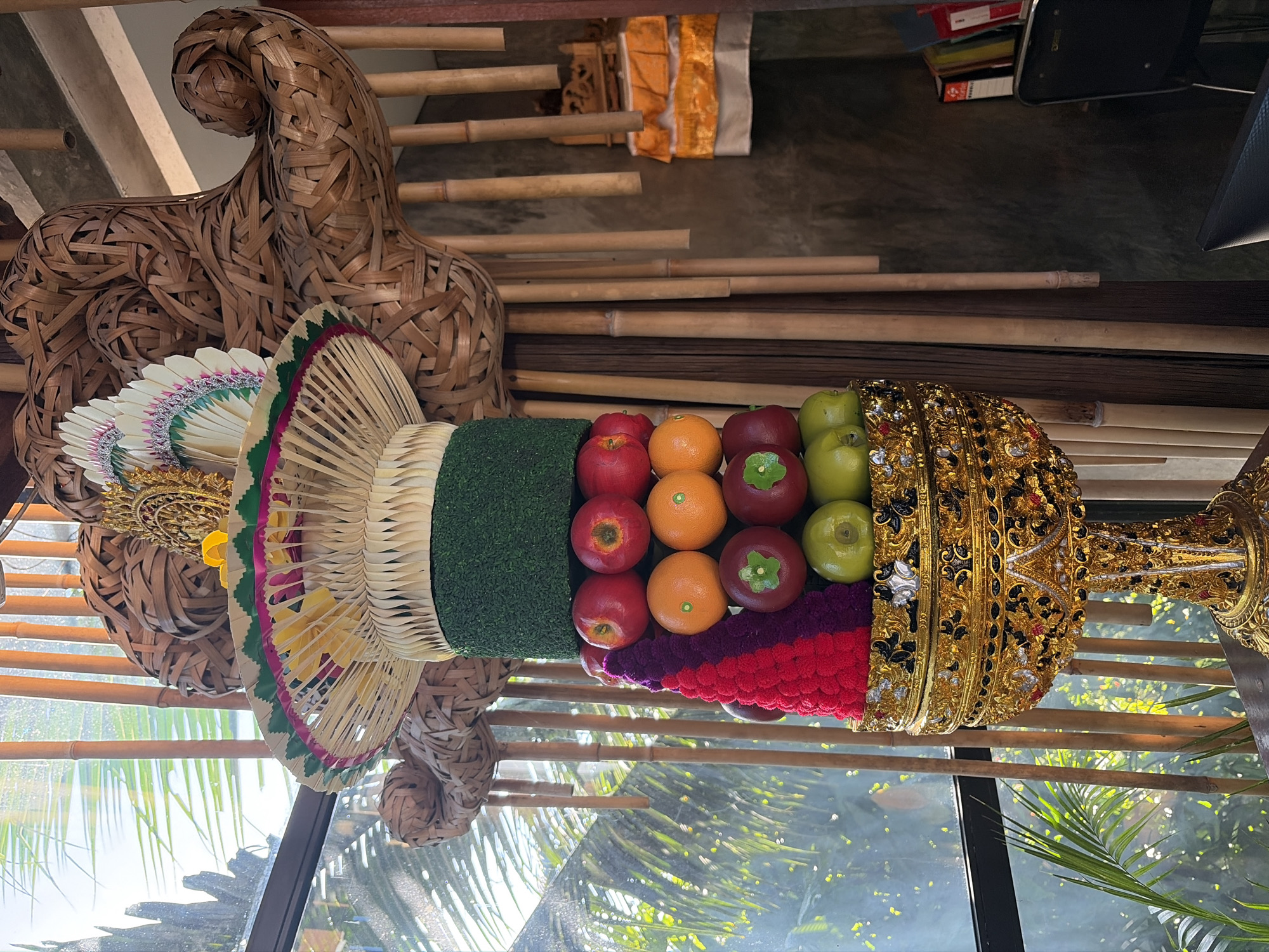 This is a vertical, medium shot of an elaborate Balinese fruit offering or "canang sari" placed in a golden, ornate stand. The offering is made up of layers of fresh fruit, including red apples, oranges, and green apples, interspersed with colorful woven materials in shades of red and purple. Above the fruit is a layer of green moss-like material, followed by a woven bamboo-like structure topped with a decorative fan made of white and green leaves and a golden emblem. The entire structure is positioned in front of a window with sunlight streaming through, illuminating palm fronds outside. Behind the offering, bamboo poles are visible, along with a small altar with orange and white fabric. The background includes a textured gray wall and a glimpse of books and a black chair on the right. The scene appears to be indoors, possibly in a restaurant or shop in the Tegallalang region of Indonesia, known for its rice terraces and traditional crafts. There are no people visible in the image.
