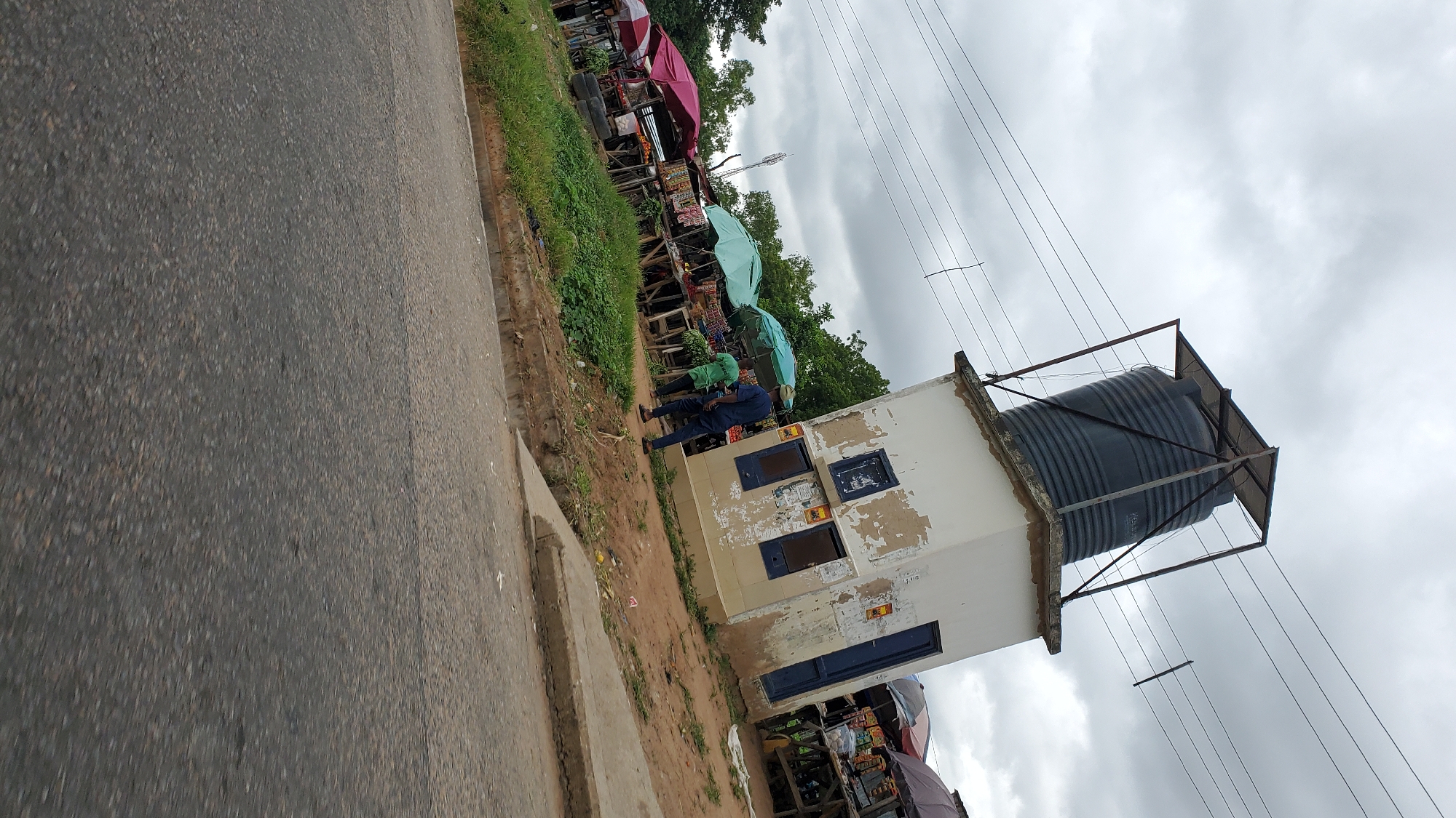 The image depicts a street scene in Jalingo, Nigeria. In the foreground, a paved road takes up the lower half of the frame. The road appears to be a typical urban street with some wear and tear. To the left of the road, a grassy embankment slopes upwards. Beyond the embankment, a row of market stalls is visible. These stalls are covered with various colored umbrellas and display goods, suggesting a bustling marketplace. Behind the stalls, lush green trees form a backdrop, indicating the presence of vegetation in the area.
Dominating the right side of the image is a peculiar structure. It's a small, white building with peeling paint, topped by a large, dark water tank supported by a metal frame. This water tank appears to be a key feature, possibly part of a municipal water system or a private water storage solution for the community. The building has dark blue doors and windows, some of which are boarded up or have metal grills.
Two individuals are present near the building. One, wearing a blue uniform, stands near the water tank structure, seemingly overseeing the area. Another person, in a green t-shirt, is standing closer to the market stalls. The overall atmosphere of the image suggests a vibrant, yet somewhat rustic, urban environment, common in many Nigerian cities. The cloudy sky indicates overcast weather conditions. The presence of overhead power lines also points to the urban setting.