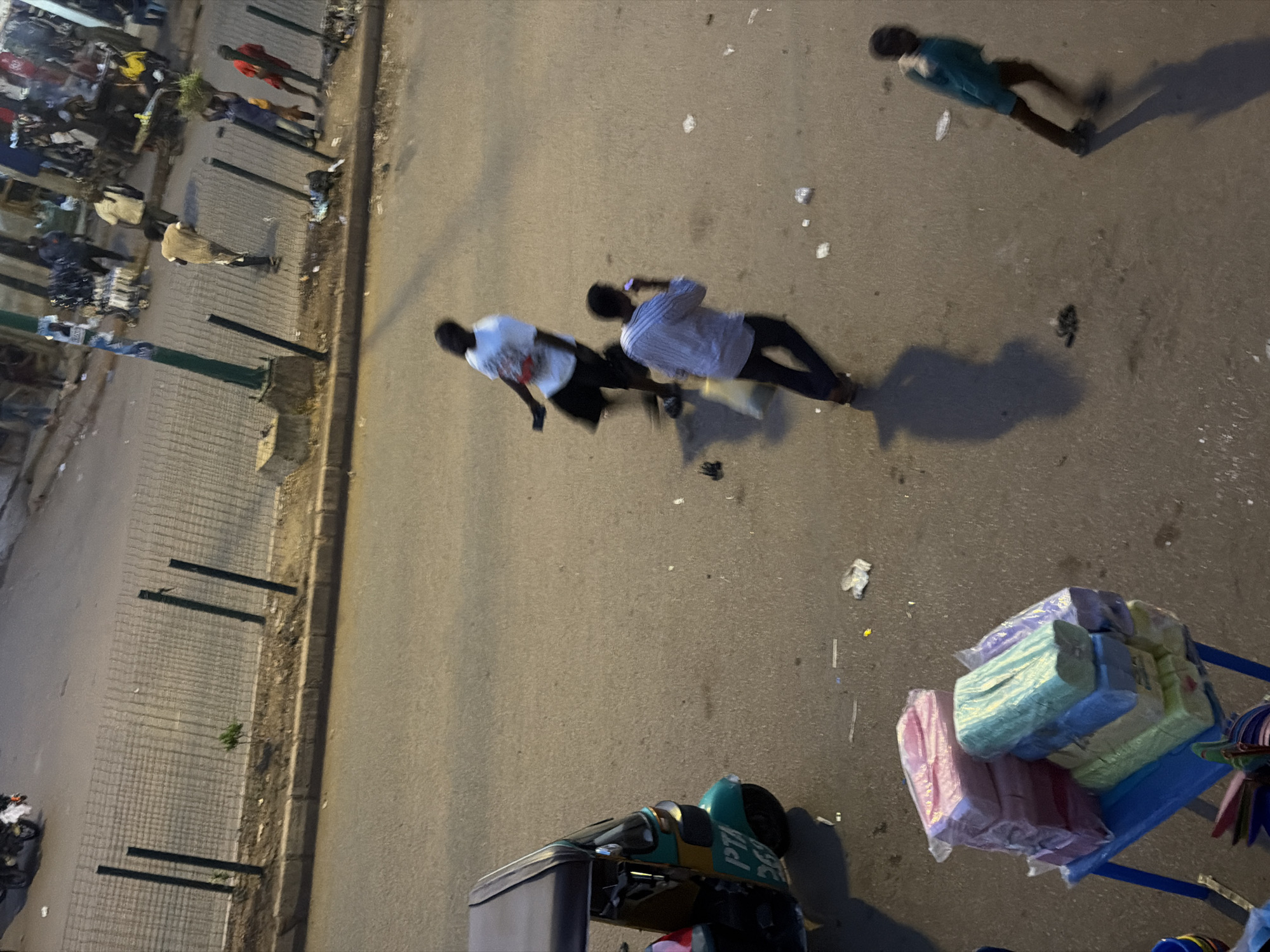 The image captures a street scene in Bukuru, Nigeria, at dusk. In the foreground, a blue plastic table is laden with stacks of packaged items, appearing to be toilet paper or similar disposable paper products in various colors, including pink, green, yellow, and purple. To the left, the rear of a green and yellow autorickshaw, identified by "PTA 269" on its side, is visible.
Further into the scene, two men are walking on the asphalt road. One man, wearing a white t-shirt with red accents and dark shorts, is in motion. The second man, dressed in a light-colored striped shirt and dark pants, is holding a cell phone to his ear. To the far right, a young boy in a turquoise sweatshirt and dark shorts is also walking, casting a long shadow.
In the background, several other individuals are present, some standing and others walking, near what appears to be market stalls or shops. A wire mesh fence runs along the edge of the road, bordering a dirt area with sparse vegetation. A motorcycle is parked further back on the road. The lighting suggests it is either early morning or late evening, with artificial lights likely contributing to the illumination. Scattered debris is visible on the road surface.