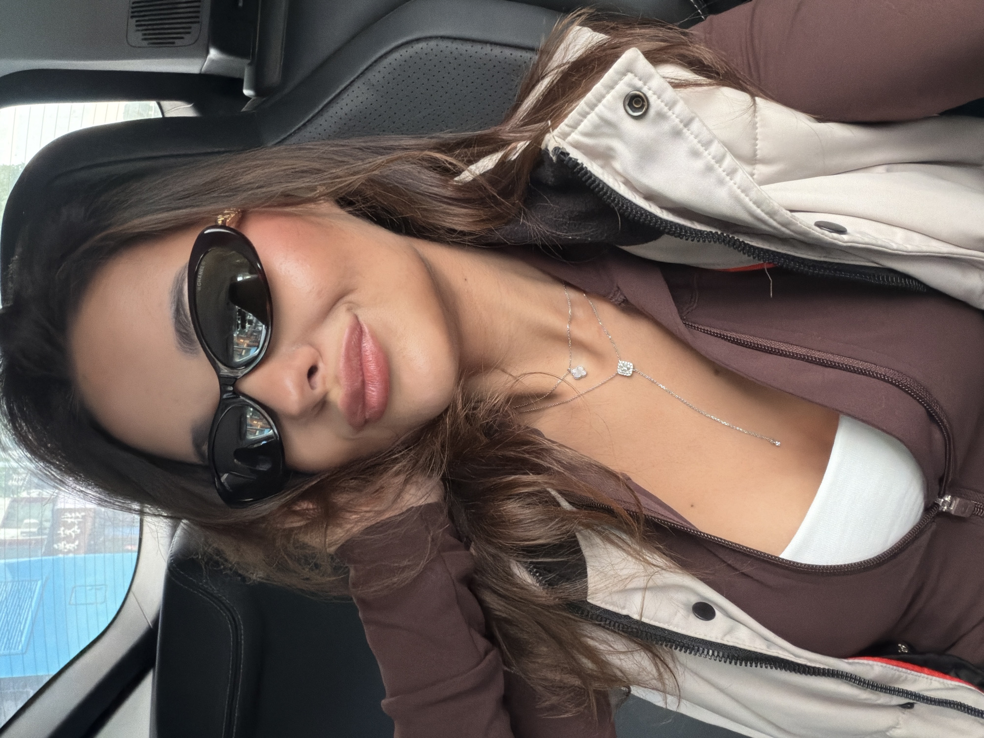 A woman wearing sunglasses, a brown jacket with a white and black vest over it, and a silver necklace is in a vehicle. The interior of the vehicle appears to have black leather seats. Outside the car window, buildings are visible.