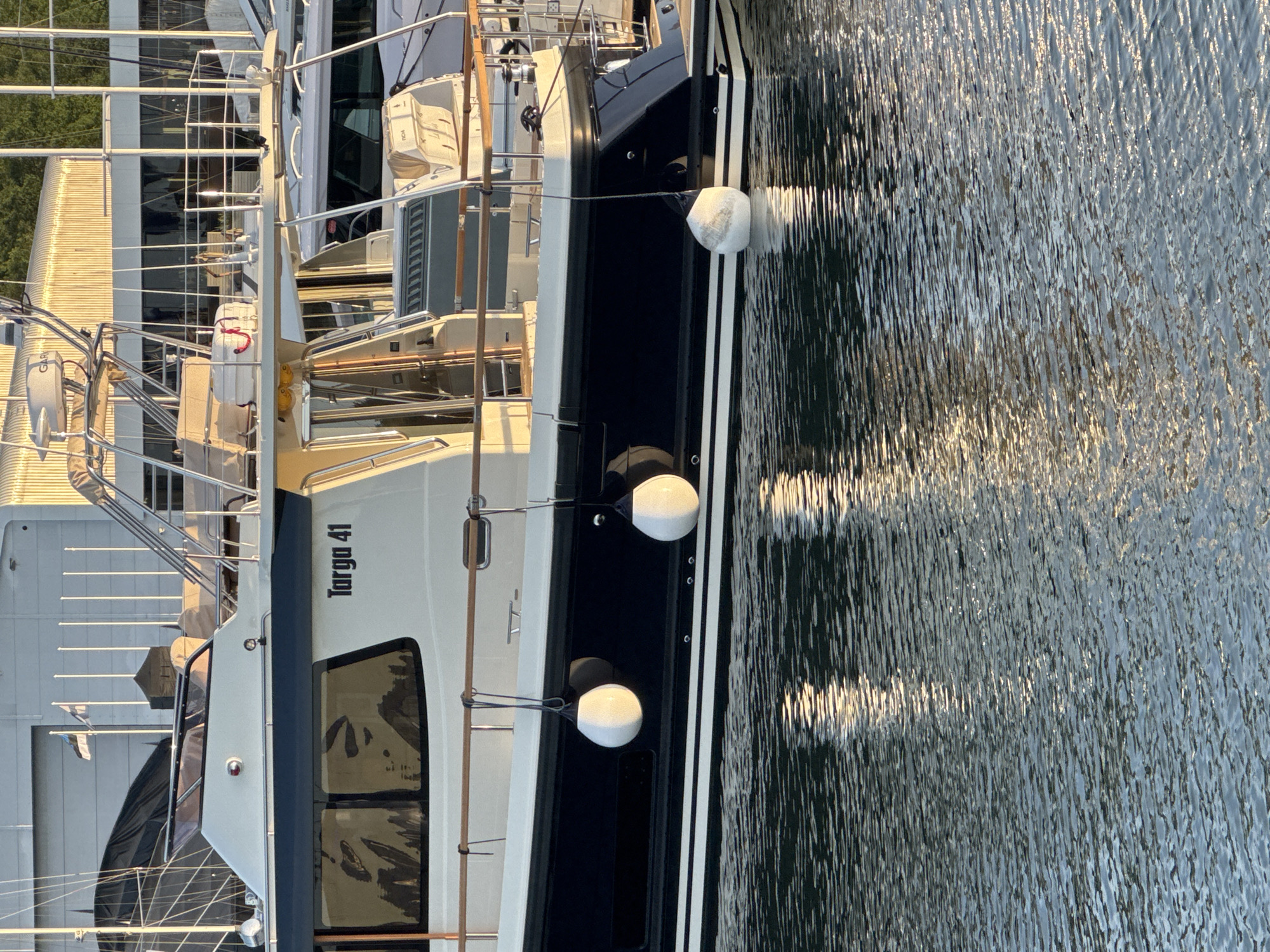 This image depicts a Targa 41 boat docked in Tallinn, Estonia, likely at a marina. The boat is white with a dark blue hull and features several white fenders attached to its side. The stern of the boat is visible, and the water surrounding it is calm with reflections of the boat and surrounding structures. In the background, a large building with a corrugated metal roof is visible, along with masts and rigging from other boats. Two flags are flying on poles in front of the building, one of which appears to be the Estonian flag. The sun is casting long shadows, indicating it is either early morning or late afternoon. No people are visible in the frame.
