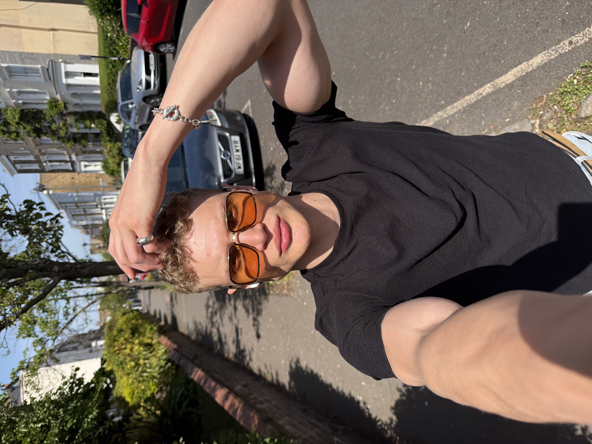 A young, light-skinned individual wearing orange-tinted sunglasses and a black shirt is taking a selfie outdoors. They are standing on a paved street lined with buildings, greenery, and parked cars. A silver bracelet adorns their raised arm, and a ring is visible on their finger. A portion of a white and beige shoe can be seen. The location is within the city of London, United Kingdom.