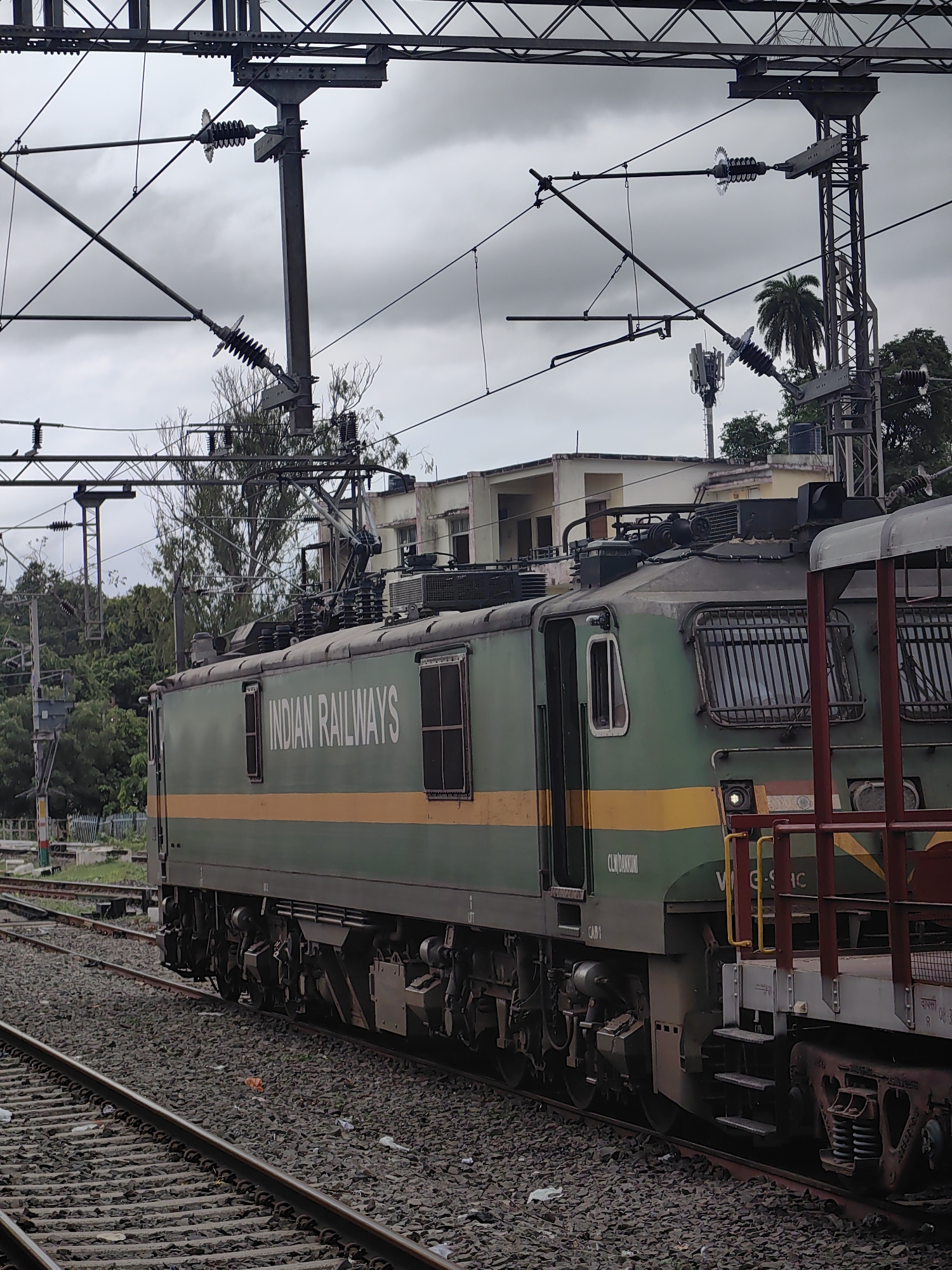 The image depicts a green and yellow Indian Railways locomotive at a train station in Khandwa, India. The locomotive is positioned on railway tracks, with multiple overhead electrical wires and supporting structures visible above. A portion of another railcar, painted red and white, is visible to the right of the locomotive. The background shows buildings, trees, and a cell tower, under a cloudy sky. The railway tracks are covered with ballast. The locomotive has the words "INDIAN RAILWAYS" printed on its side in white lettering. A portion of the locomotive's identification number, "WAG-9IC", is visible. The image is taken from a slightly elevated perspective, looking towards the side of the train.