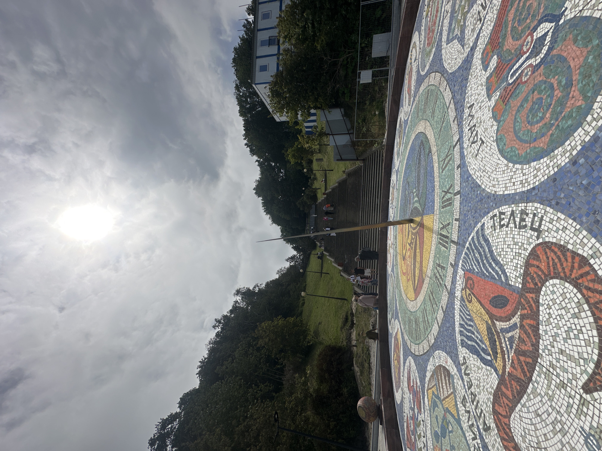 In Svetlogorsk, Russia, a mosaic sundial, featuring zodiac symbols and Roman numerals, is prominently displayed in the foreground. A tall gnomon stands at the center of the sundial. In the middle ground, a wide staircase ascends a grassy hill, lined with trees. Several people are seen walking up the stairs. In the background, a building with blue stripes on a white facade is visible atop the hill, surrounded by dense foliage. The sky is overcast with bright sunlight filtering through the clouds.
