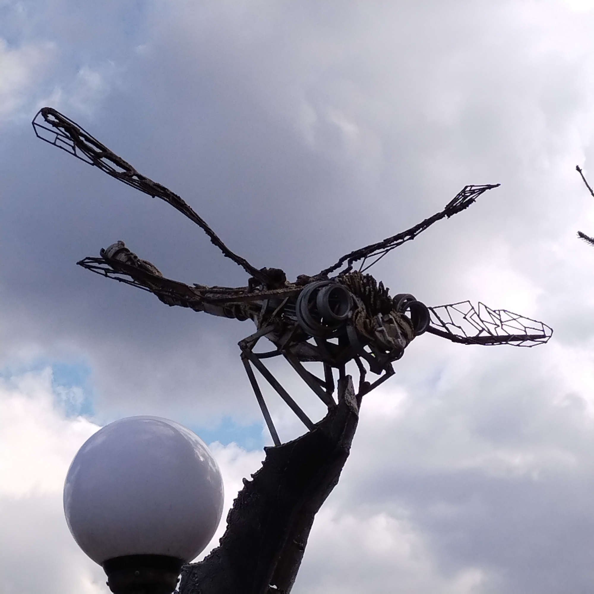 The image shows a metal sculpture of a dragonfly, likely made of repurposed metal parts. The sculpture is perched atop a rough, trunk-like base next to a globe light fixture. The sky in the background is overcast, with grey and white clouds. The sculpture is located in the city of Nizhny Tagil, Russia.