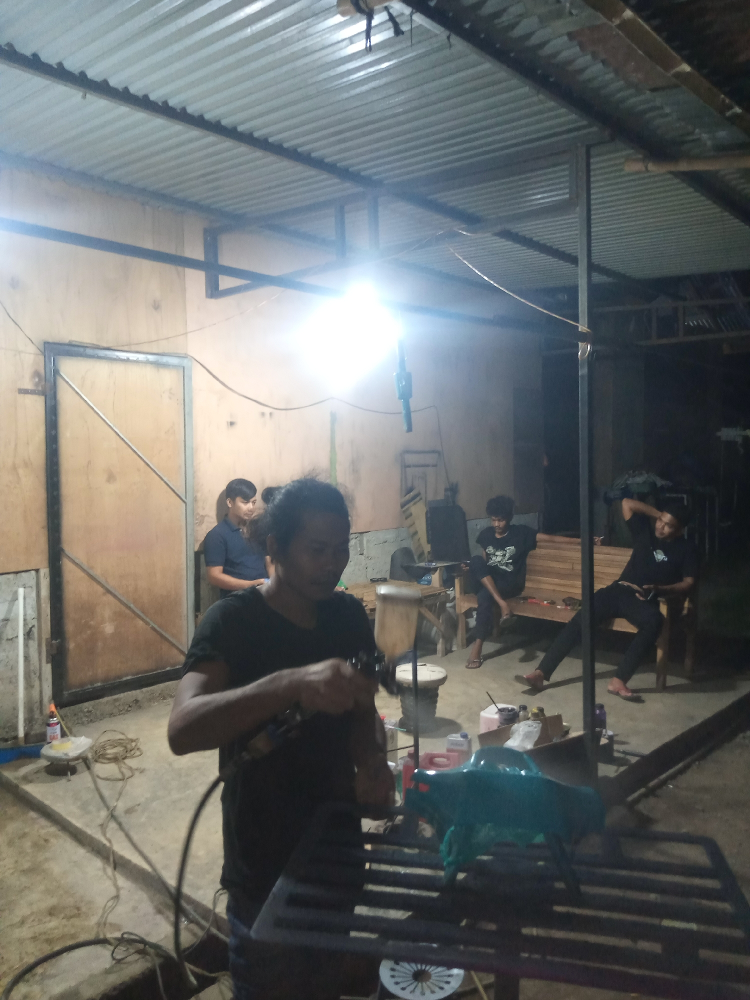 The image depicts an indoor workshop or garage, possibly located in Balee Rastong, Indonesia, given the context. The space is dimly lit, with a bright overhead light illuminating the central area. The ceiling is made of corrugated metal, supported by exposed metal beams. The walls appear to be constructed of wood panels.
In the foreground, a person wearing a dark t-shirt is holding what appears to be a spray gun and is actively engaged in work. Their hands and the tool are the main focus of their attention.
Behind the person in the foreground, several other individuals are present. To the left, a person in a dark blue shirt stands near a large wooden door or gate. Further back, two individuals are seated on a wooden bench. One is wearing a dark t-shirt and jeans, while the other is in a black t-shirt and dark pants, with their arm resting behind their head. Another person in a black t-shirt is seated on a wooden bench to the right of the image, looking towards the left.
Various objects are scattered around the workshop floor, including electrical cords, containers, and what looks like a teal-colored object resting on a metal grate in the mid-ground. The floor is concrete. The overall impression is of a functional workspace where multiple people are present, possibly working on or supervising a project.