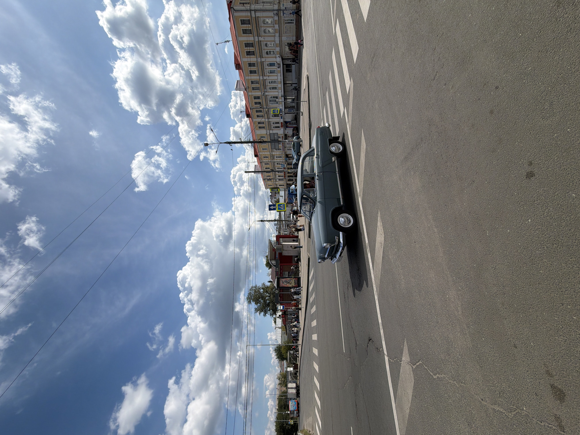 A vintage gray sedan is driving across a crosswalk on a wide asphalt road in the city of Nizhny Novgorod, Russia. The car is angled towards the right side of the frame and its front fender is closest to the viewer. The sky above is a vibrant blue with scattered white, puffy clouds. In the background, there are multi-story buildings with yellow facades and red roofs, a bus stop, and a crowd of people. Power lines are visible stretching across the sky. The scene appears to be a sunny day in an urban environment.