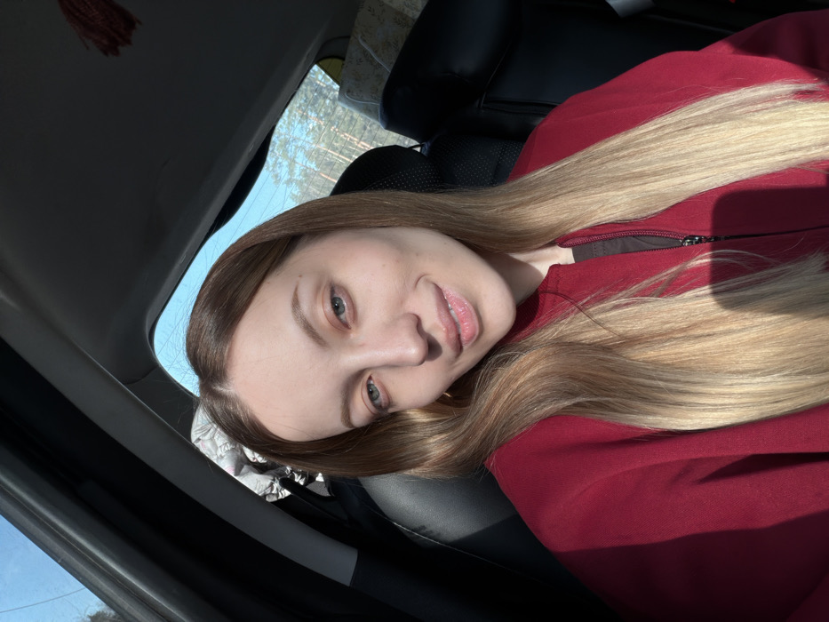 A female individual is captured in a close-up selfie from inside a vehicle, located in Selevino, Russia. She possesses long, straight light brown hair with prominent blonde highlights at the ends and light-colored eyes. Her attire consists of a dark red zip-up jacket worn over a dark inner layer. Her lips are slightly parted in a subtle smile, and she is looking directly into the camera. The vehicle's interior features dark-colored seats and panels. Sunlight enters the vehicle, illuminating the subject's right side and casting shadows on the left. Through the rear window, a dense forest composed of tall trees is visible under a clear blue sky. Within the car's interior, a crumpled light-colored garment is visible in the rear on the viewer's left, and a patterned fabric item rests on the rear seat on the viewer's right. A small, red, fibrous object hangs from the rear ceiling of the vehicle.