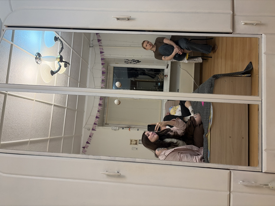 A mirrored wardrobe reflects a bedroom scene. The top mirror shows a person sitting on a chair near a window, while the ceiling light is visible. The lower mirror reflects a person lying on a bed taking a picture with a phone. Decorations and bedding can also be seen in the mirrors. The wardrobe itself is white with silver handles.