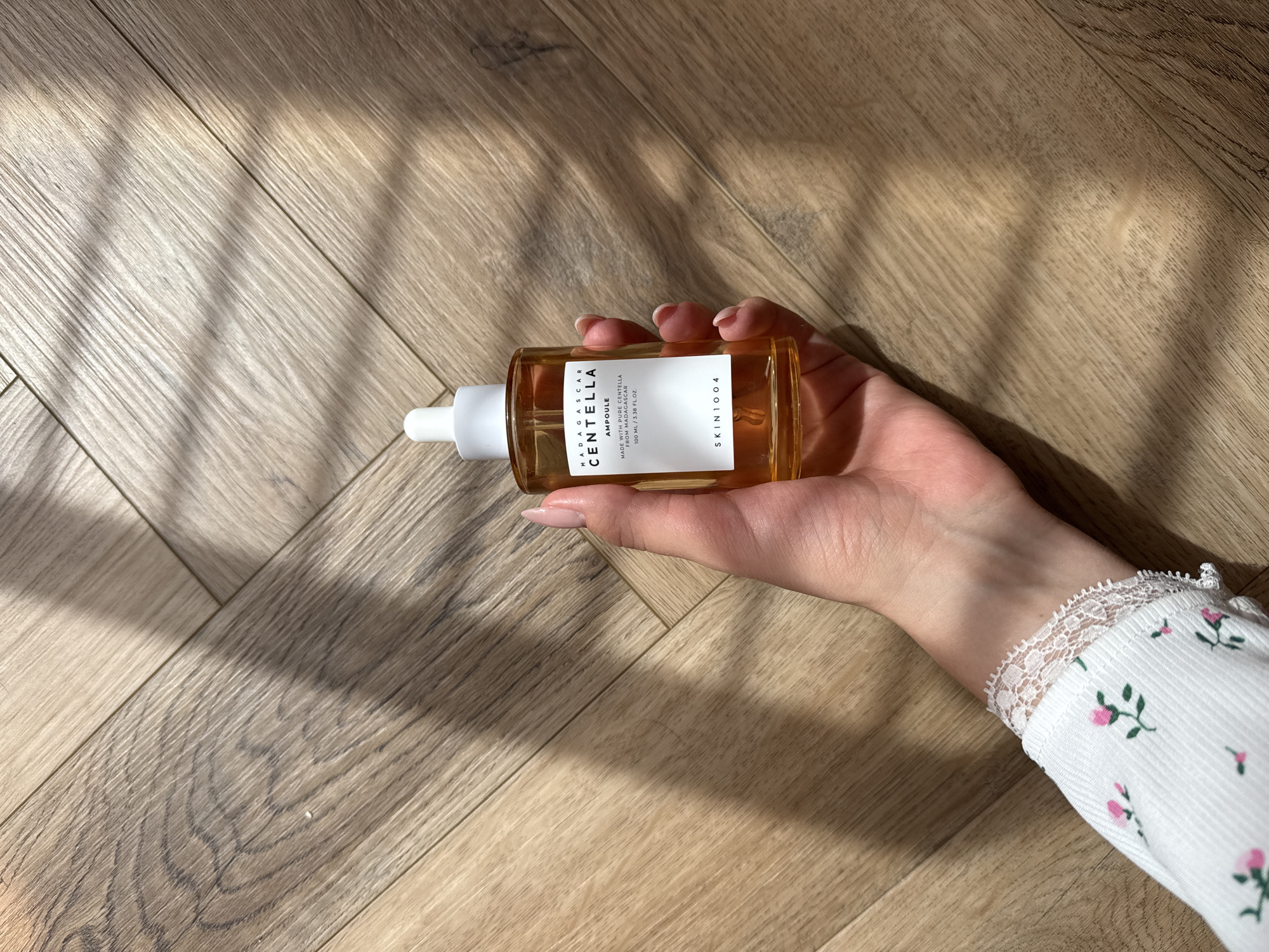 A hand holds a bottle of Madagascar Centella Ampoule by SKIN1004 in a wooden floor environment with sunlight. The bottle is amber-colored with a white dropper and label with black text that reads "MADAGASCAR CENTELLA AMPOULE MADE WITH PURE CENTELLA FROM MADAGASCAR 100 ML / 3.38 FL. OZ. SKIN1004". The hand has manicured nails and is part of an arm wearing a white long-sleeved shirt with a floral pattern and lace trim. The wooden floor has a herringbone pattern with visible wood grain and is partially illuminated by sunlight casting shadows across the surface.