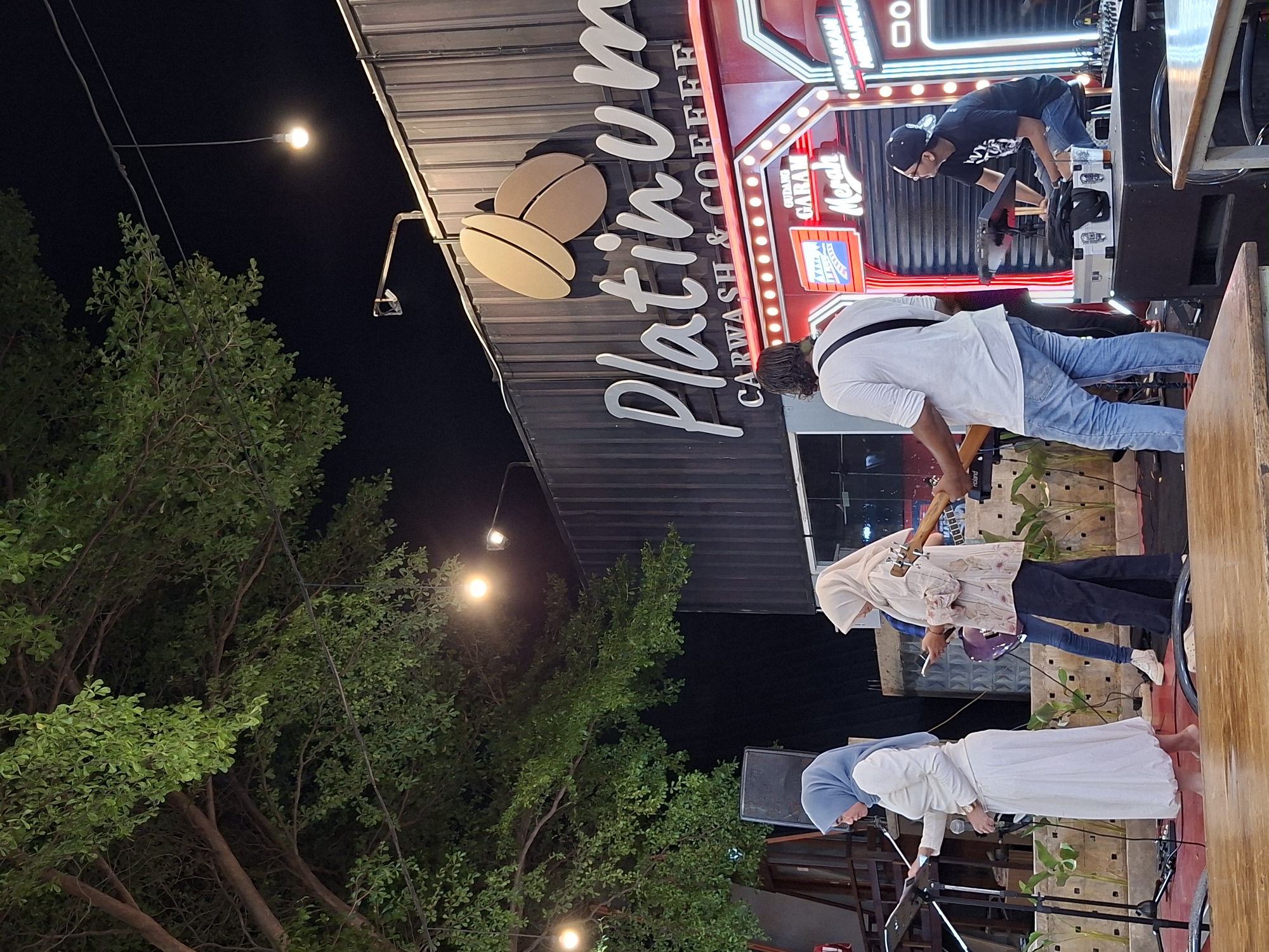 A dimly lit outdoor night scene in Lhokseumawe, Indonesia, features a live musical performance at "Platinum Carwash & Coffee," signified by a prominent neon sign. A band of four people performs on a raised platform. The band consists of a person playing keyboard/synthesizer, a guitarist, and two vocalists wearing white and blue hijabs, respectively. A sound engineer or technician is situated at the rear of the stage. The surrounding environment is partly obscured by a large tree and overhead streetlights.