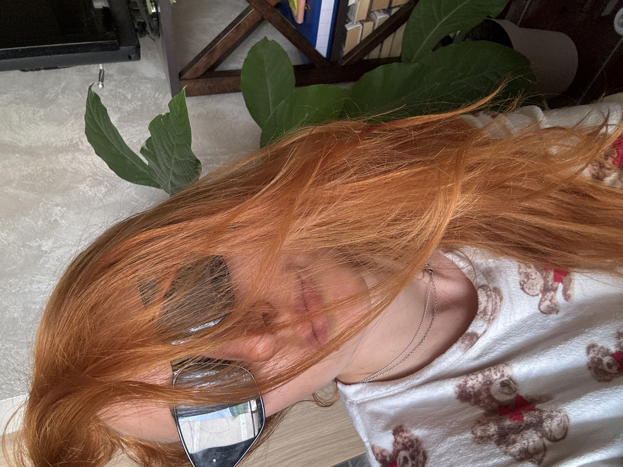 A person with red hair is lying on a surface covered in a teddy bear-patterned fabric. The person's face is partially obscured by their hair and a pair of sunglasses. In the background, green leaves are visible, along with elements that appear to be part of a room interior, potentially a TV on a piece of furniture. There's no explicit indication of the location being Budva, Montenegro.