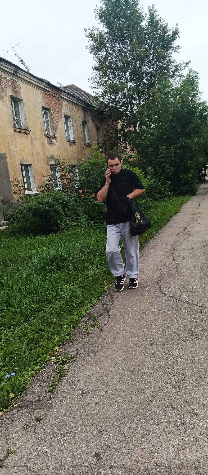 A man with short brown hair and a beard is walking on a paved path next to a grassy area. He is wearing a black short-sleeved shirt with a textured pattern and light gray sweatpants. He is carrying a black bag over his right shoulder and is holding a black mobile phone to his left ear. He is looking towards the camera with a neutral expression.
Behind the man is a dilapidated, light yellow building with multiple windows, some of which are boarded up or appear broken. The building shows signs of decay, with exposed brickwork. Dense green foliage and trees are also visible behind the man and to the right of the building. The path is cracked and shows wear. The sky is overcast.
This scene is located in the city of Izhevsk, Russia, specifically on an unpaved road in front of an old residential building. The specific address is unclear, but it appears to be a residential area.