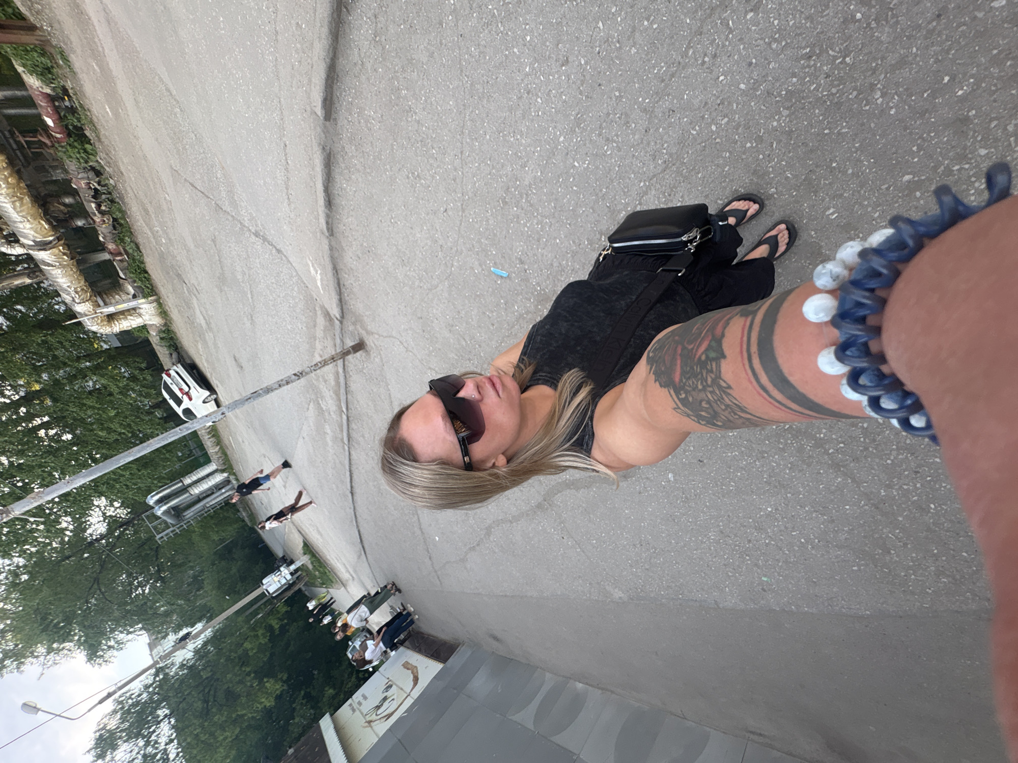 A top-down, medium shot captures a woman with blonde hair and sunglasses wearing a black tank top and black sandals, standing on a paved street. She has a black crossbody bag and a tattoo on her arm. A blue hair tie with white beads is wrapped around her wrist closest to the camera.
In the background, there is a glimpse of a city street scene with trees, buildings, and vehicles. A group of people is walking in the distance. The location appears to be a street in Perm, Russia, with pipes and trees on the left.