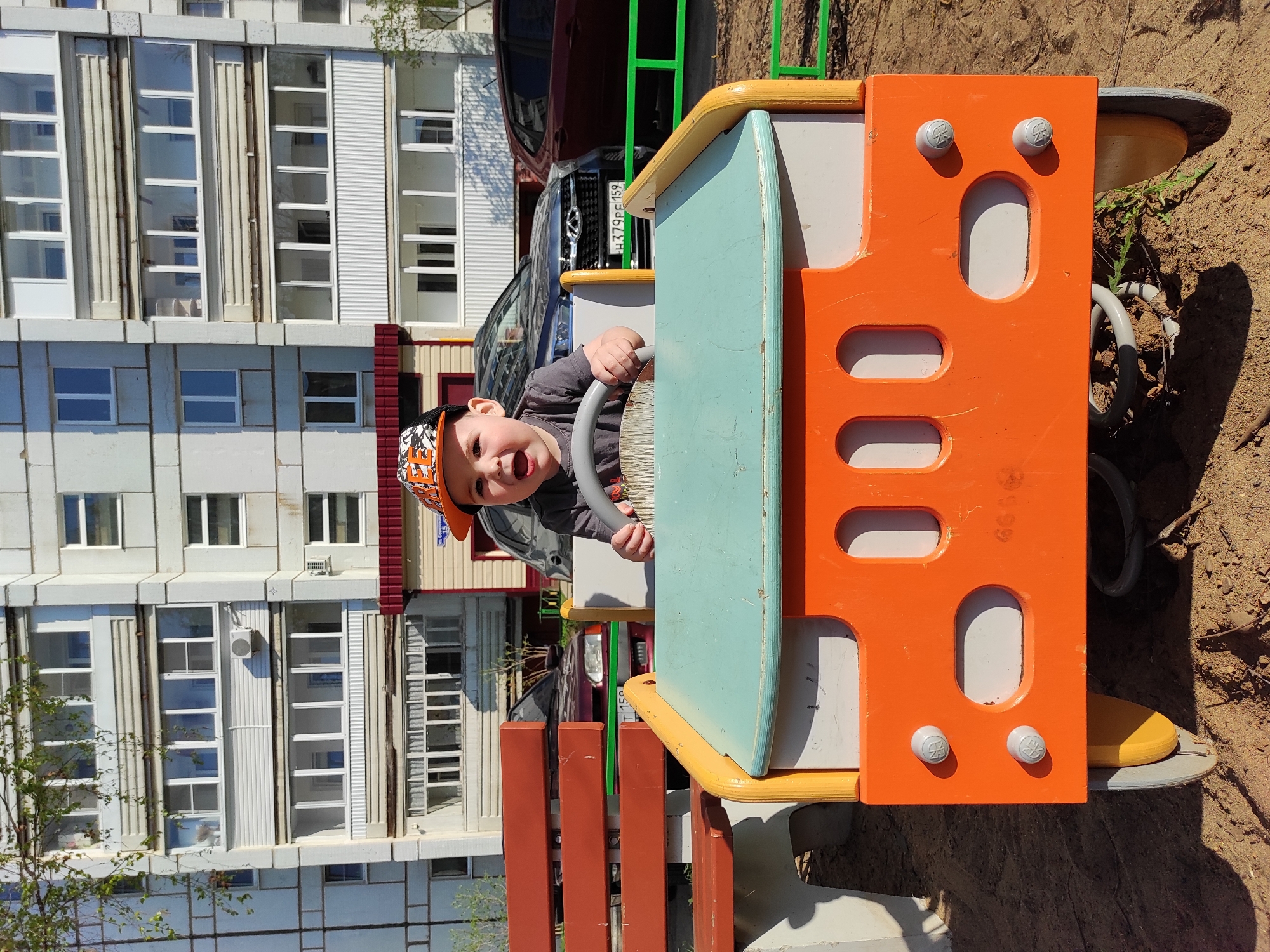 A young child, approximately 1-2 years old, is playing on a colorful spring rider in what appears to be a playground. The child is wearing a cap with orange and black designs and a grey shirt. The spring rider is orange, light blue, yellow, and white, with cut-out shapes. Behind the rider is a residential building with multiple windows. A car is parked in front of the building. The location is identified as Chaikovsky, Russia.