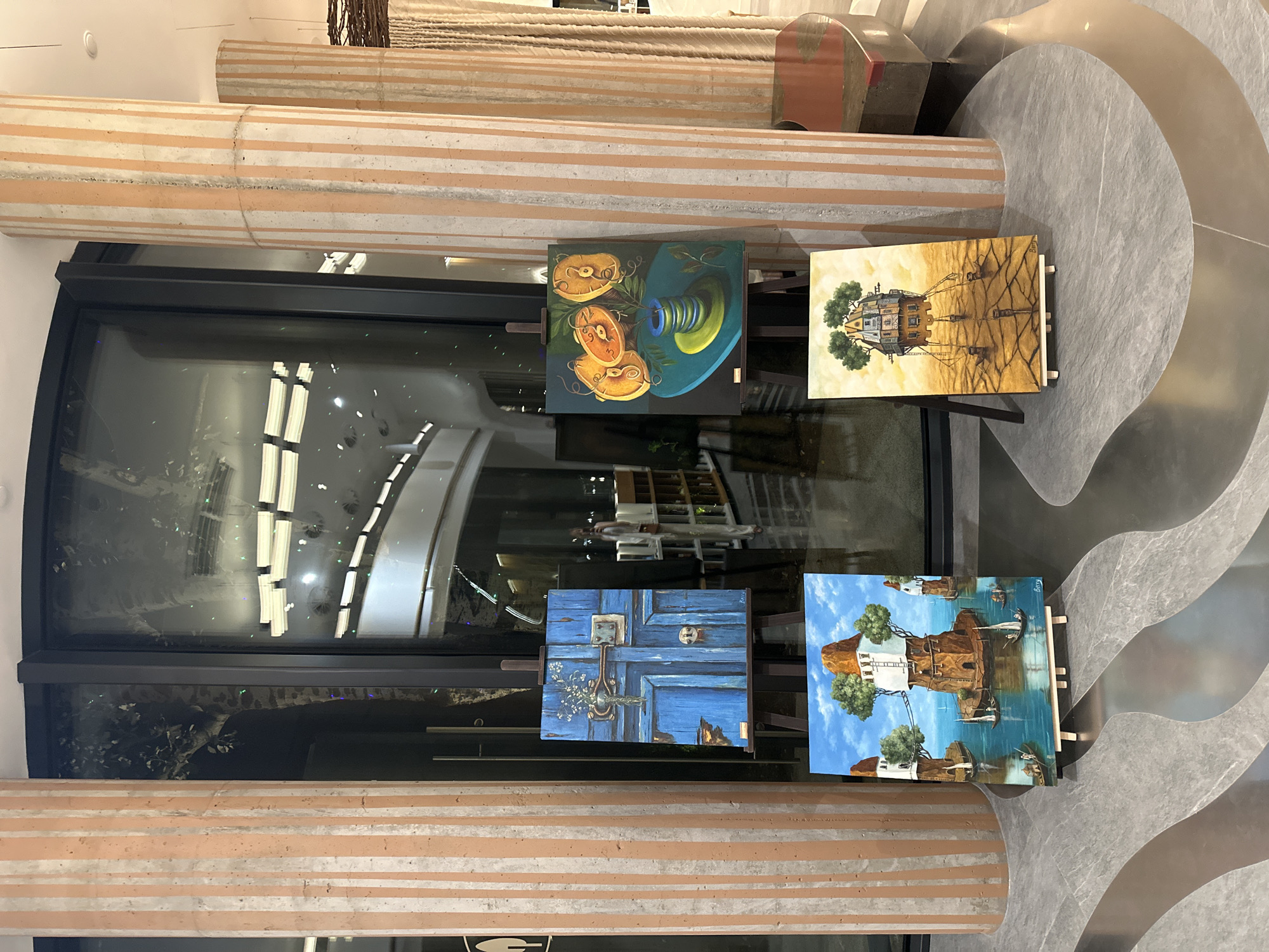 The image shows four paintings displayed on easels in an indoor space. Two large, fluted columns with orange and white stripes are prominent on either side of the scene, framing a large glass window or door. The floor is a polished, dark grey material with a wavy, lighter grey pattern.
From left to right, the paintings are:
1. A close-up of a weathered, bright blue door with a brass lock and handle, and a small bouquet of white flowers above the lock.
2. A still life featuring three orange slices with seeds and a spool of blue thread on a teal surface.
3. A surreal landscape with houses built on stilts in a body of water, connected by bridges, with sailboats. The sky is blue with white clouds.
4. A surreal landscape depicting a multi-story house on tall stilts, standing on cracked, golden-brown earth under a hazy sky.
A person, possibly a woman, can be seen reflected in the glass window in the background, appearing to be in a library or shop with bookshelves. A curved, modern bench with a reddish-brown seat is visible in the far right of the image.
The location is identified as Gelendzhik, Russia. The scene appears to be an art exhibition or a display in a public space like a hotel lobby or gallery.