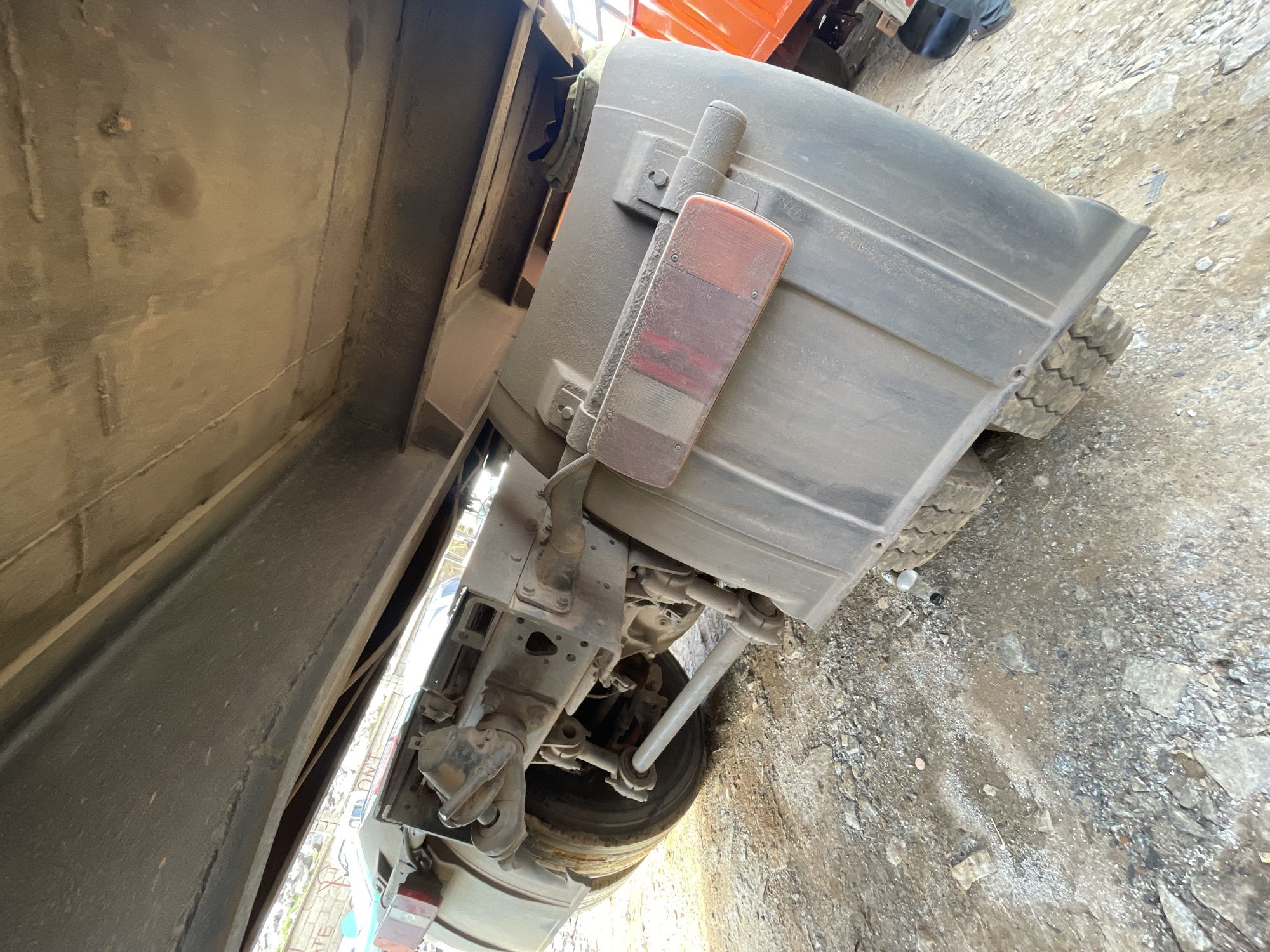 The image shows the underside and rear of a truck parked on a rough, rocky surface. The rear wheel, suspension components, and a taillight are visible. A portion of the truck's cargo bed and surrounding structures are also in view. The environment consists of a dirt and stone-covered ground. There is no information in the picture that would allow any indication about the location being Abuja, Nigeria, or any mention of people.
