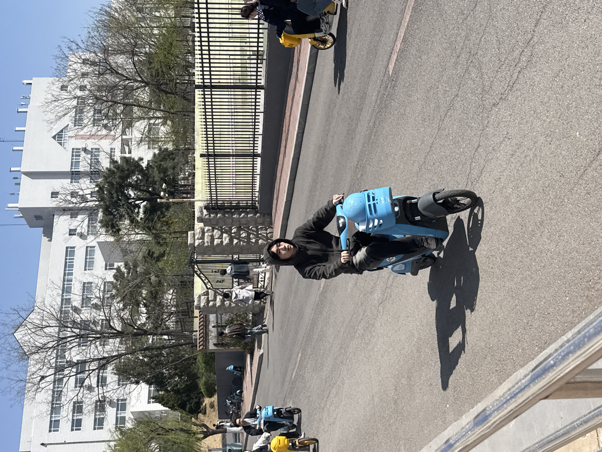 The image captures a street scene in what appears to be Zhongtun, China, based on architectural styles and objects in the image. The primary subject is an individual riding a light-blue shared electric scooter on an asphalt road. The rider is wearing dark clothing, including what looks like a hooded jacket.
Behind the rider, a wrought-iron fence separates the road from a building complex with white facade, which appears to be the campus building of the university. A group of people can be seen walking on the sidewalk behind the fence, some appearing to head towards a gate or entrance. Other similar scooters, in yellow and blue, are parked in the distance. The weather seems clear and sunny, casting shadows on the road.