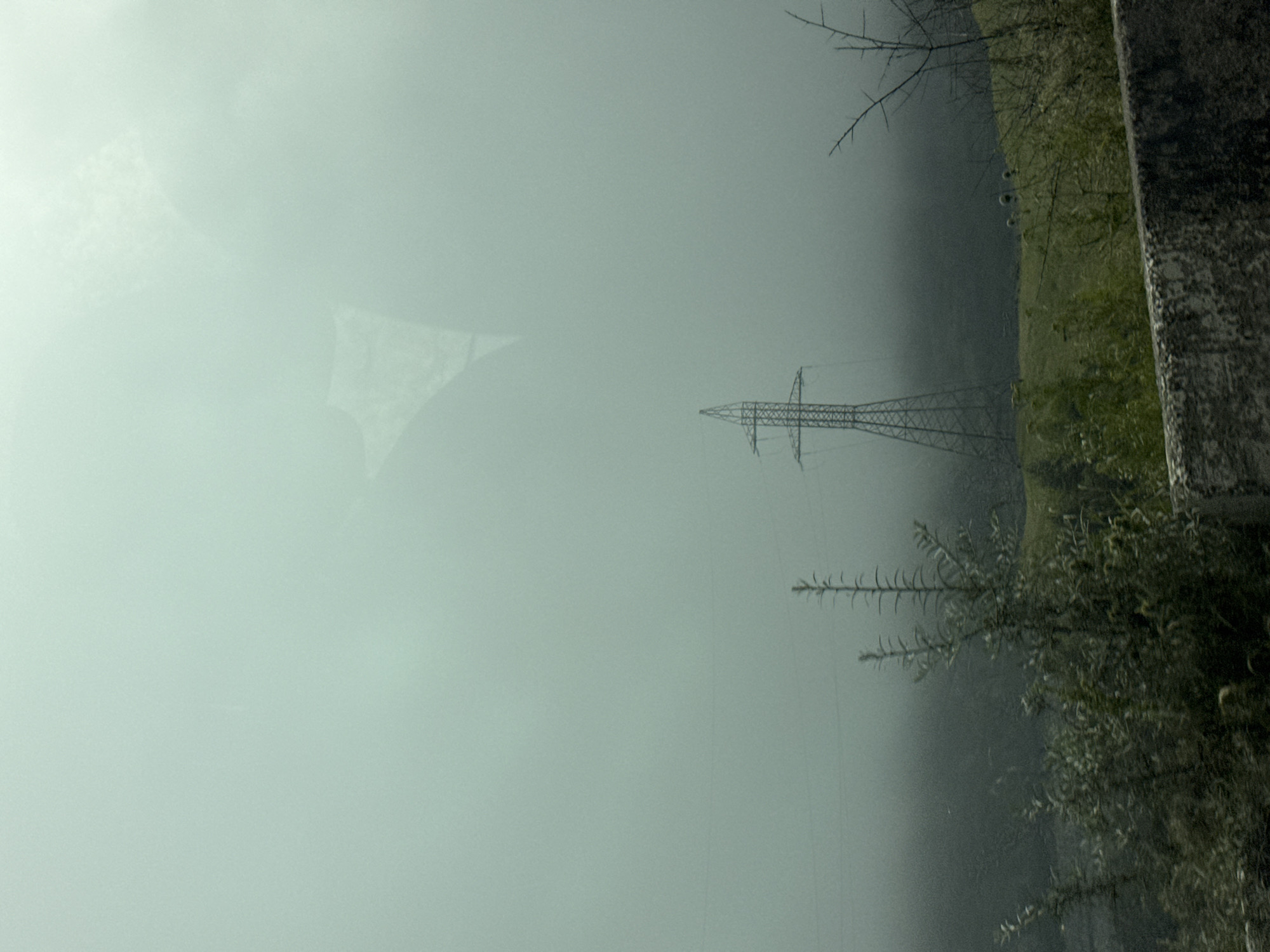 An overcast, hazy day in the city of Tsdo, Georgia. A transmission tower stands in the mid-ground, shrouded in mist, on a grassy hillside. In the foreground, a low stone wall runs horizontally across the image, its rough texture a contrast to the soft, diffused light. Sparse vegetation, including some thorny branches, grows along the wall. The sky is a pale, uniform gray, with a faint, blurry reflection of an object, possibly a person or a vehicle's interior, visible in the upper portion of the image. The overall atmosphere is one of subdued light and a sense of distance, with the natural landscape and man-made infrastructure merging into the fog.