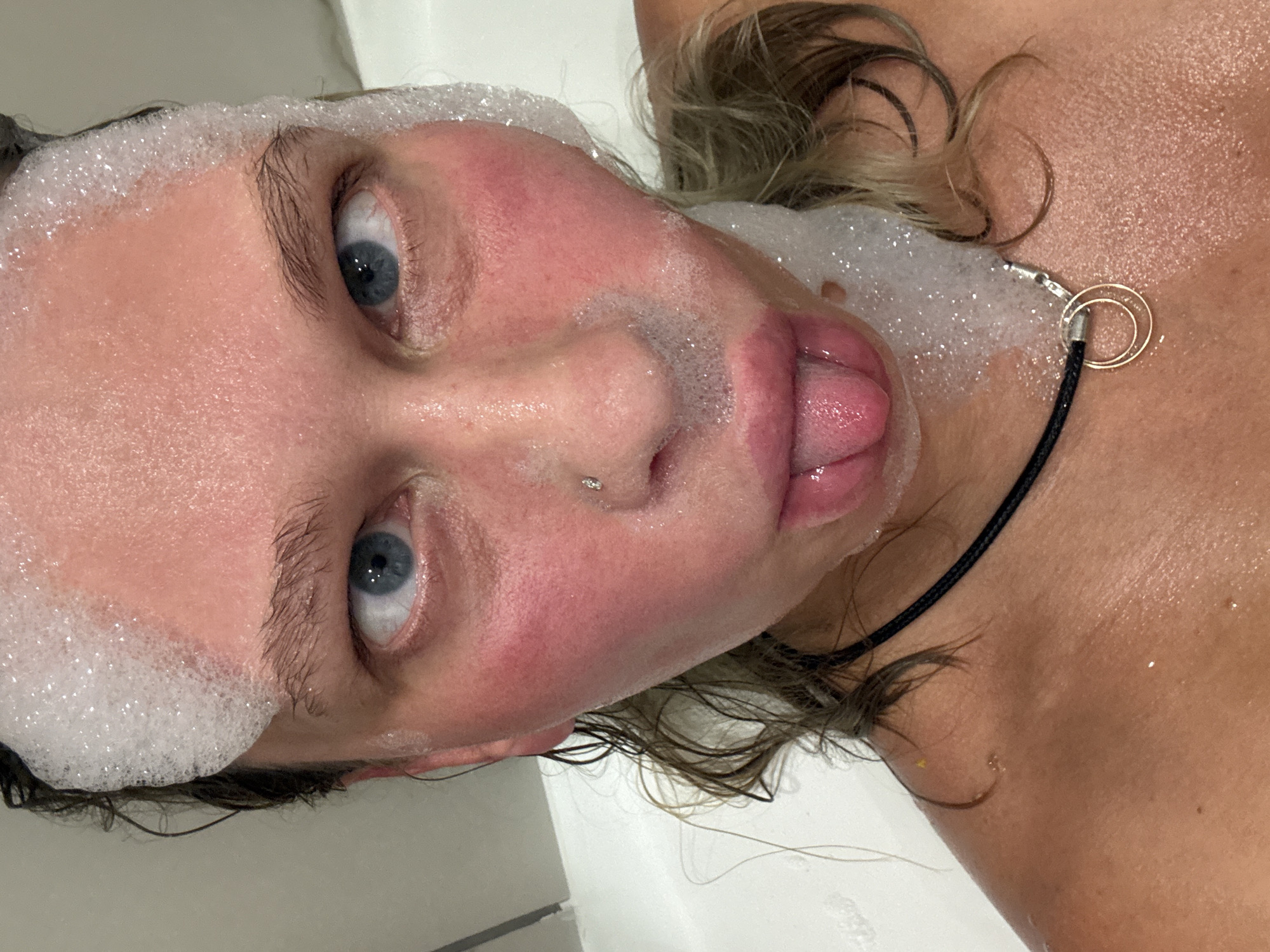 The image shows a close-up, rotated view of a person, likely a woman, from an overhead perspective, with their head and upper chest visible. The person is looking directly at the camera with wide, blue eyes. Their tongue is slightly extended, resting on or just below their lower lip, contributing to a playful or expressive demeanor.
The individual is in a setting that appears to be a bathroom, possibly a bathtub or shower, indicated by the abundance of white soap bubbles covering their forehead, hairline, and parts of their face and neck. Their skin is wet and appears flushed in areas like the cheeks and nose. They have a small, silver-colored stud piercing on their nose. Their hair, which is light-colored (blonde or light brown) and curly or wavy, is wet and partially covered by bubbles.
A black cord necklace with a distinctive golden-colored double-ring pendant is visible on their chest. The background is uniformly white, suggesting a clean, tiled surface or the inside of a tub. There is no visible text in the image. The scene is situated in Perm, Russia.